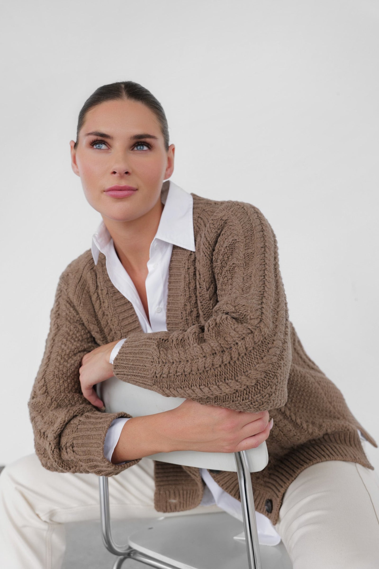 Woman wearing a brown knitted cardigan over a white shirt and pants, sitting on a chair against a white background.