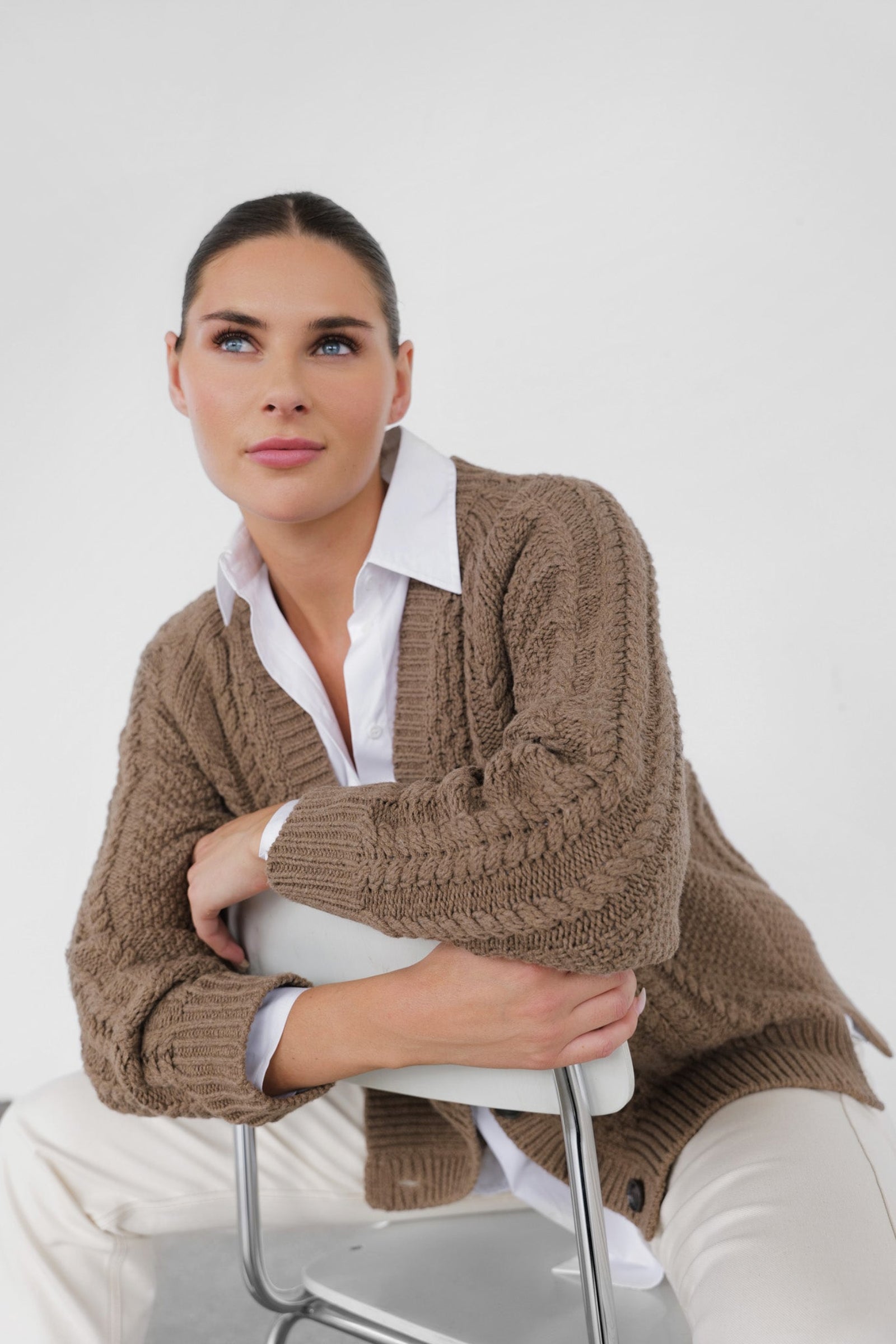 Woman wearing a brown knitted cardigan over a white shirt and pants, sitting on a chair against a white background.