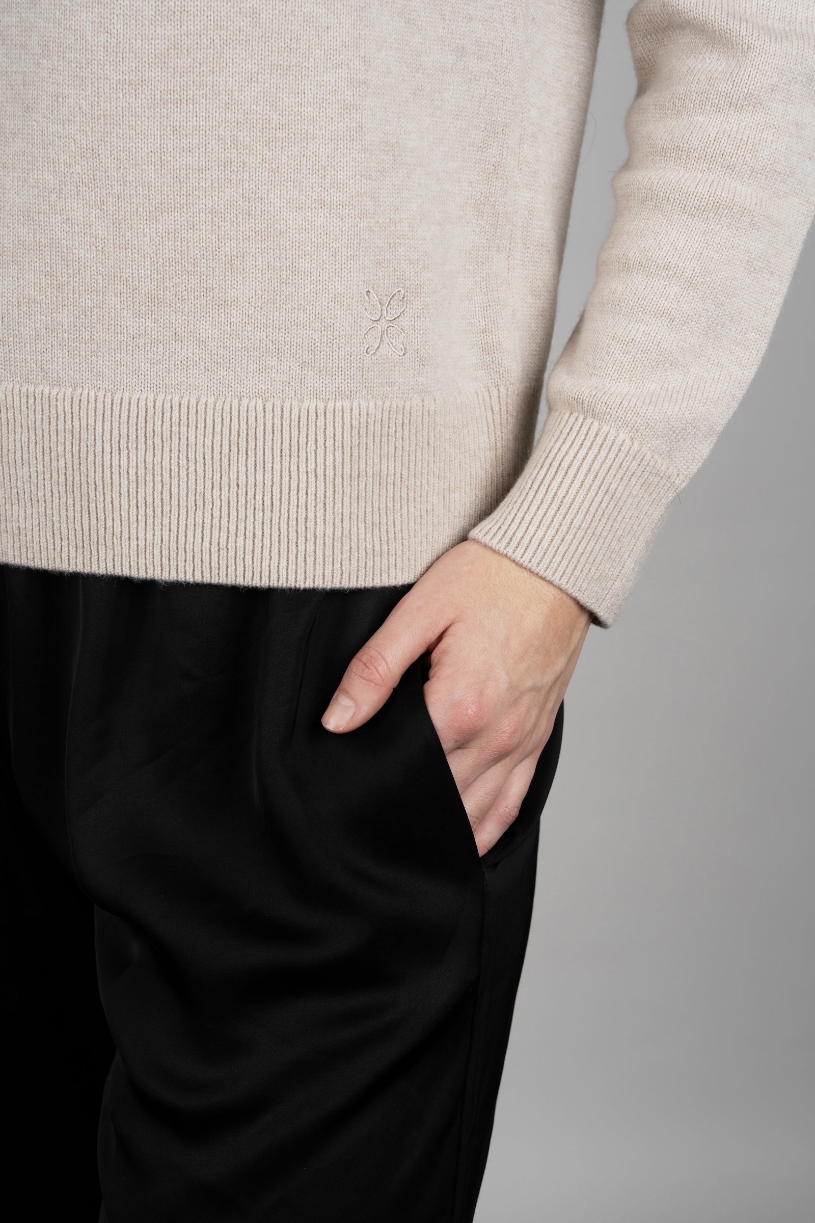 Person wearing a beige sweater and black pants against a gray background