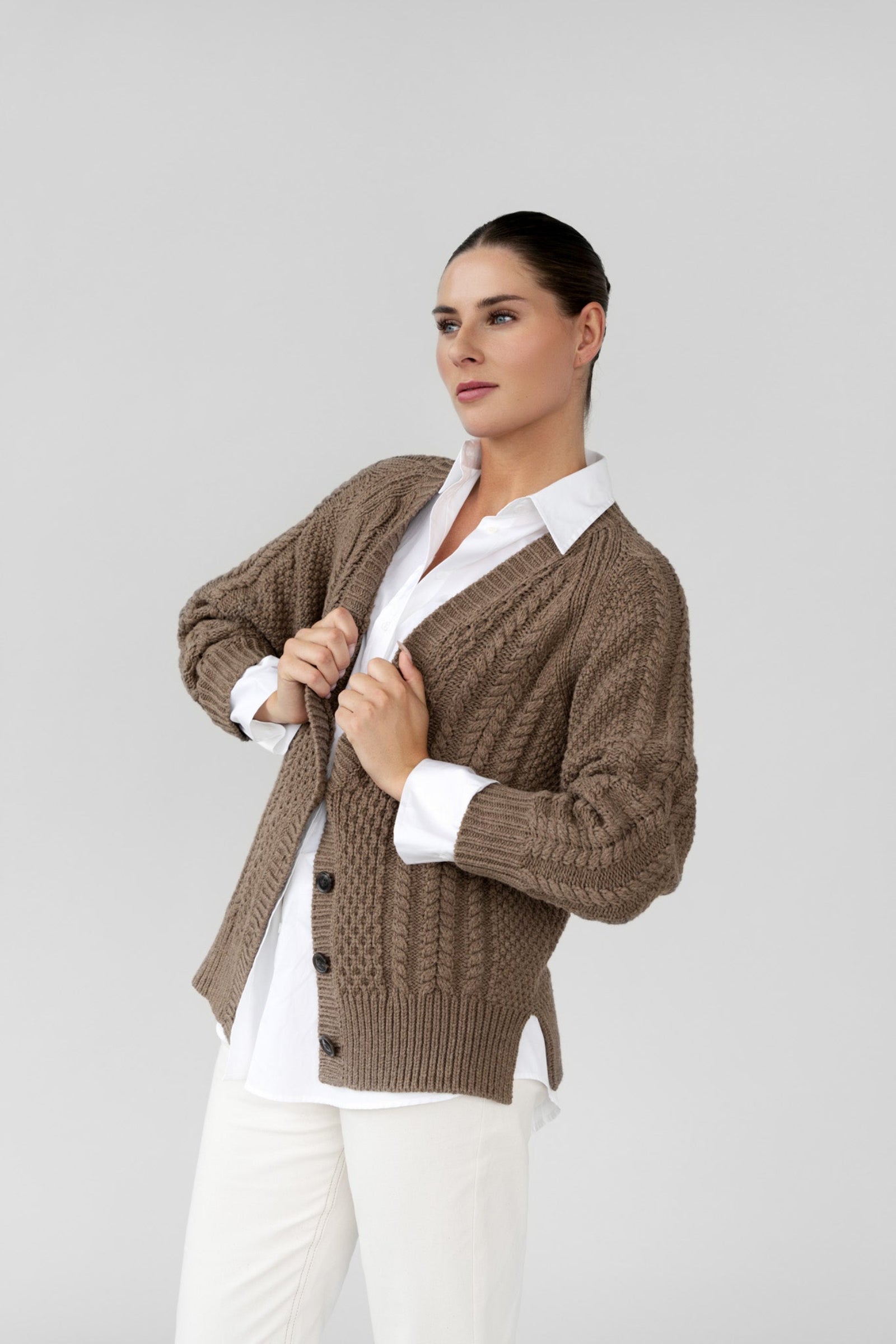 Woman wearing a brown cardigan over a white shirt and pants on a gray background
