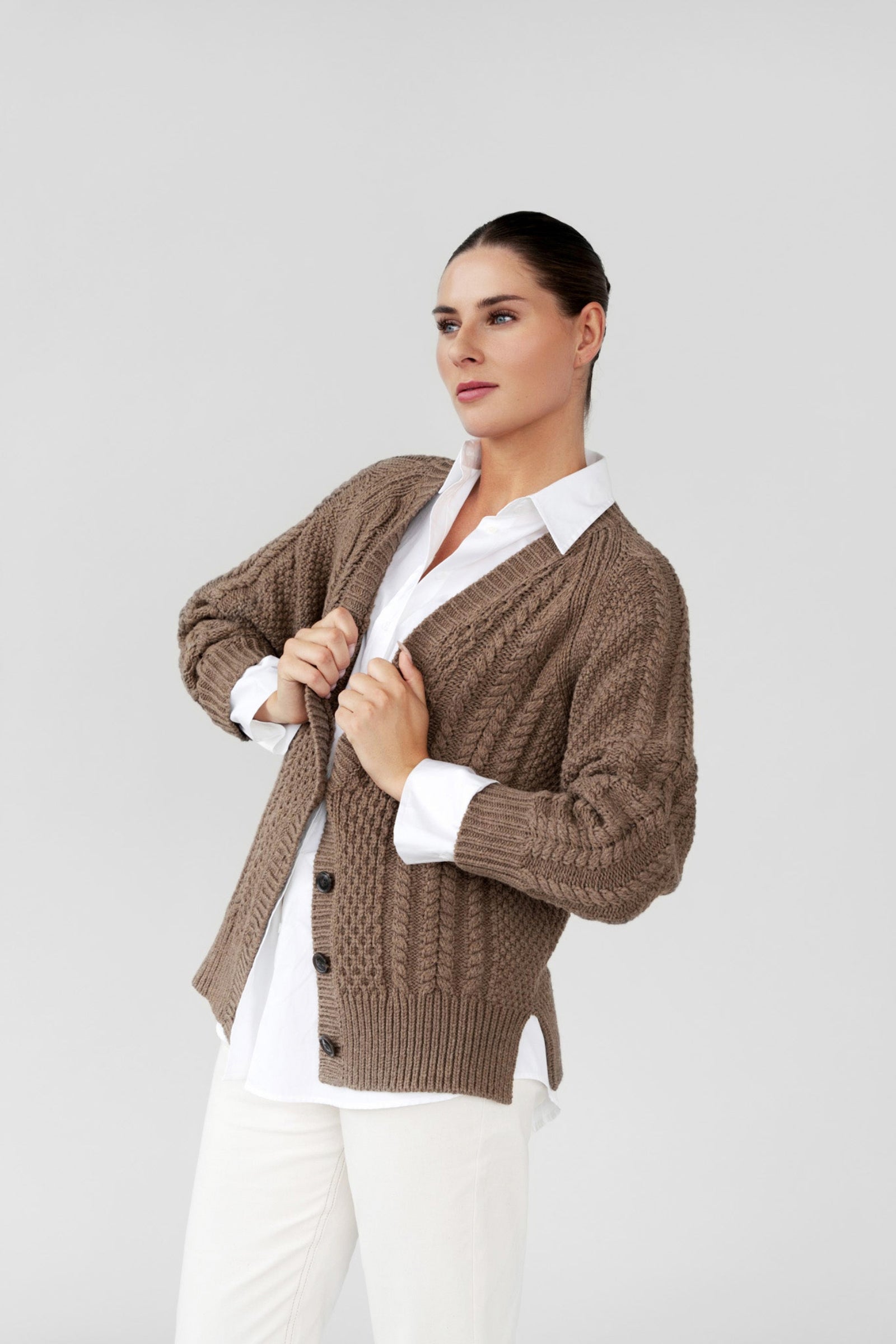 Woman wearing a brown cardigan over a white shirt and pants on a gray background