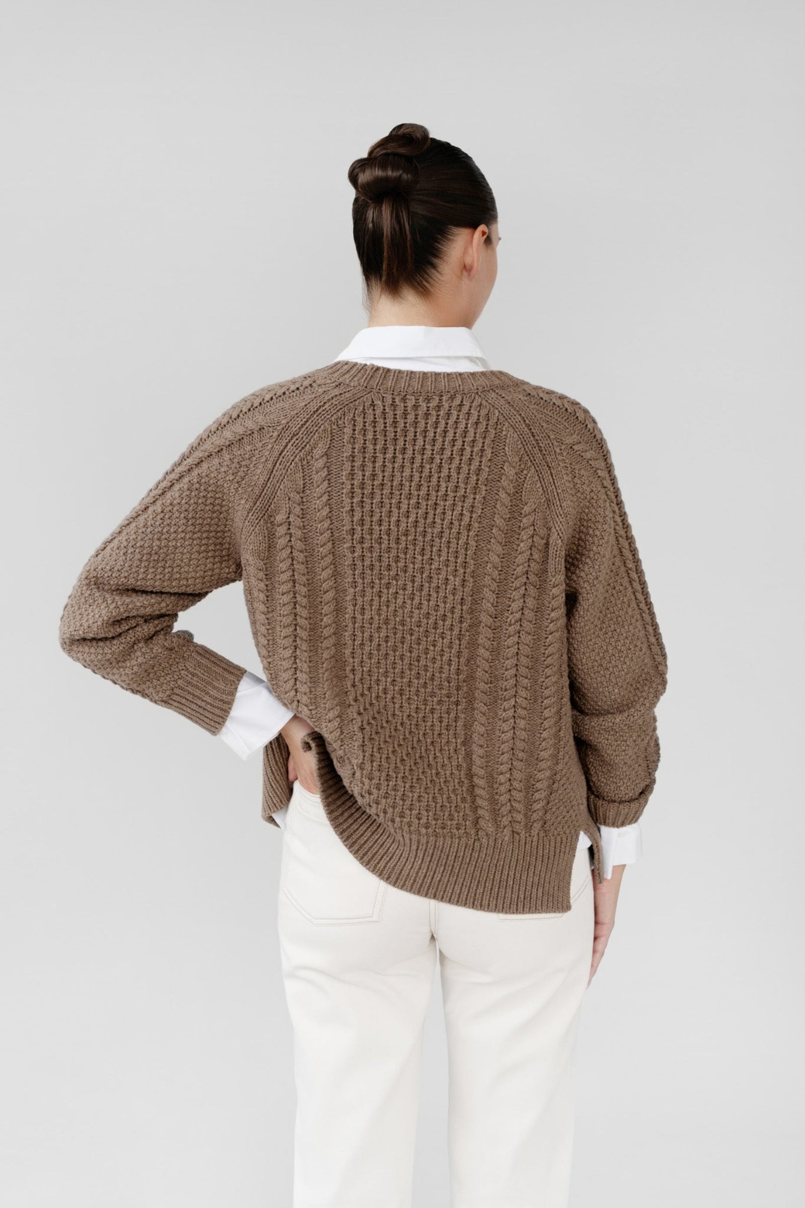 Person wearing a brown knitted sweater over a white shirt and beige pants on a plain background