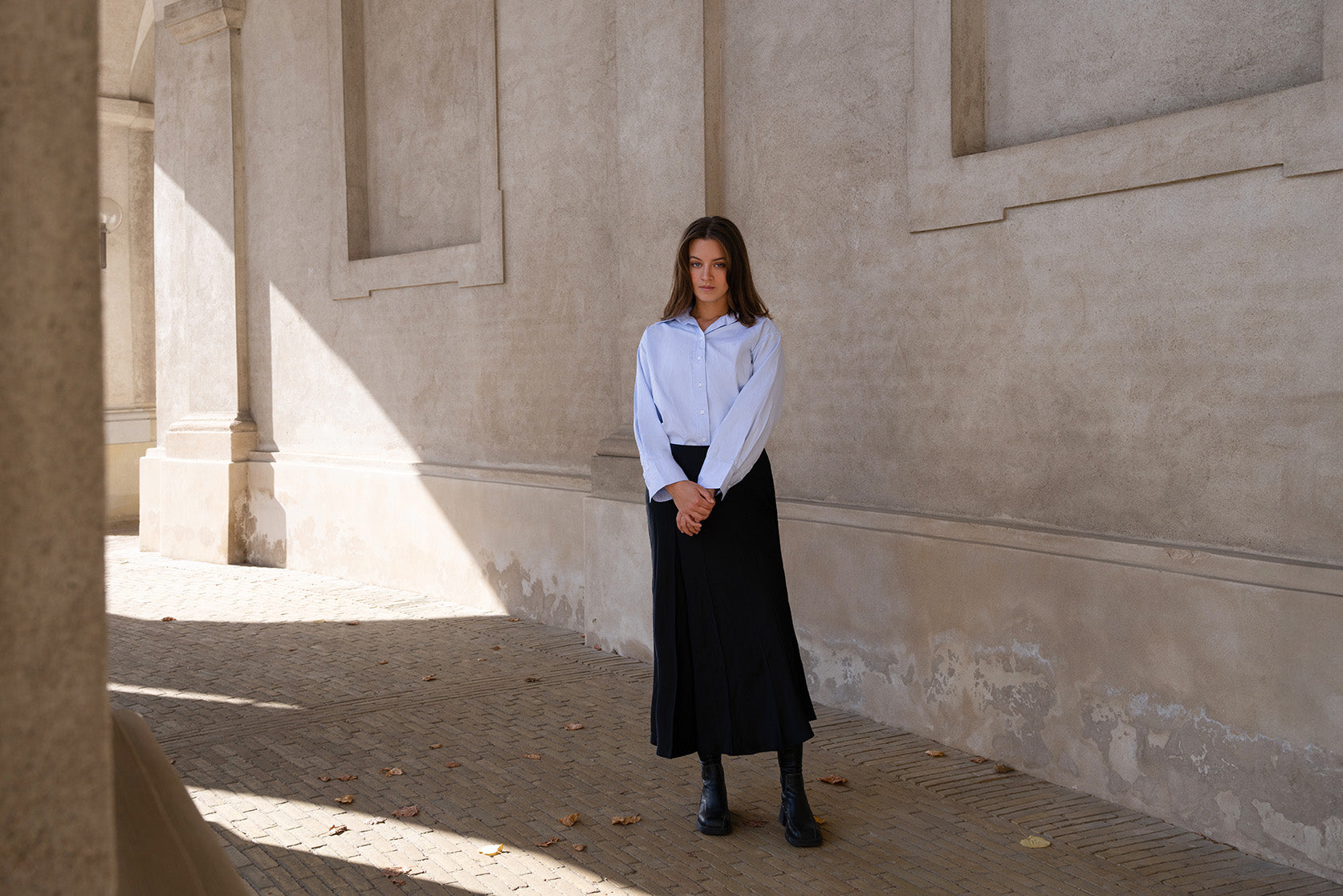 Woman in a white shirt and black skirt standing in a sunlit hallway with stone walls.