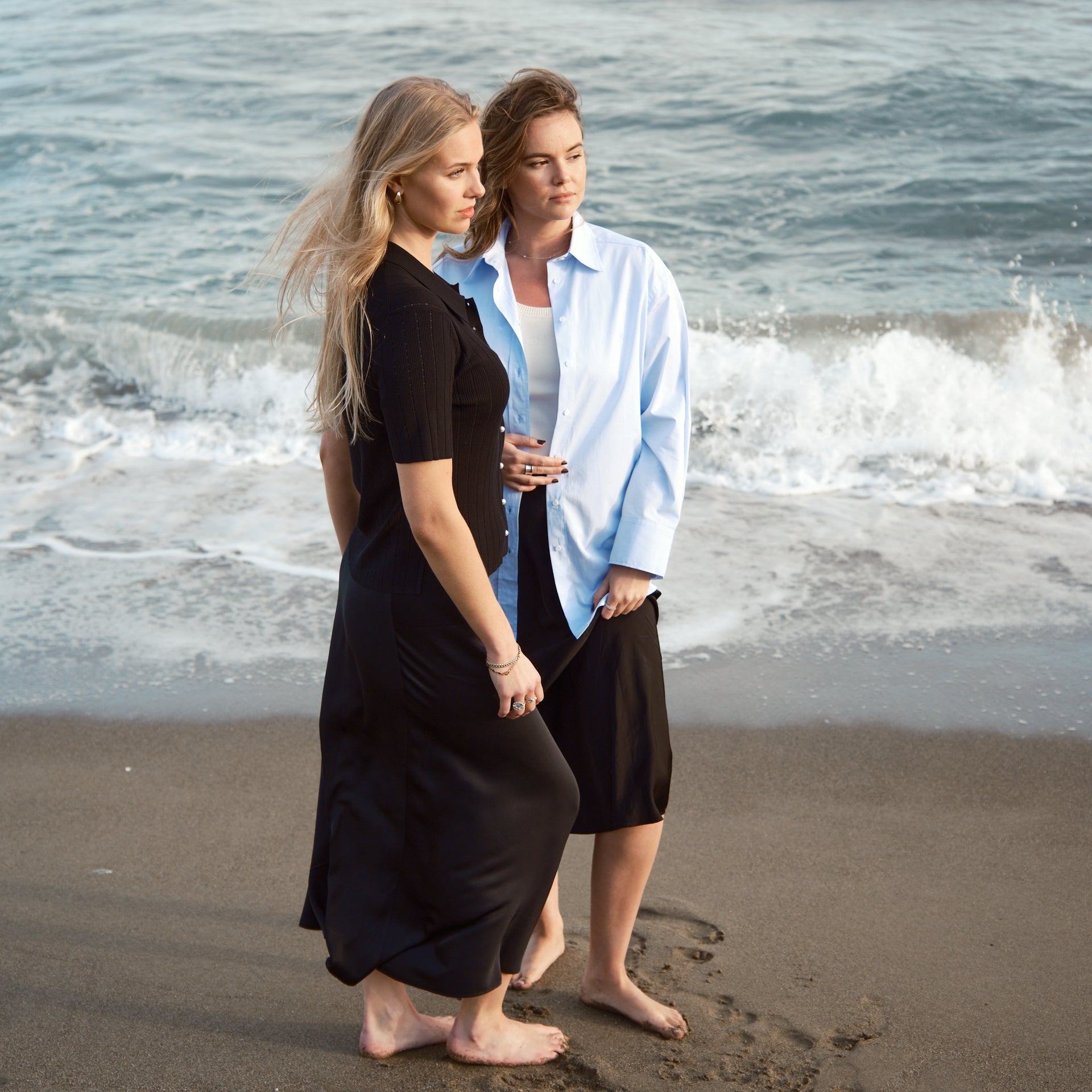 To modeller står barfodet på stranden iført CYME Copenhagen designs: den ene i sort ribstrikket cardigan og silkeskørt, den anden i blå bomuldsskjorte, hvid ribtanktop og sort silkebuks. Billedet fanger en rolig og naturlig stemning ved kysten.