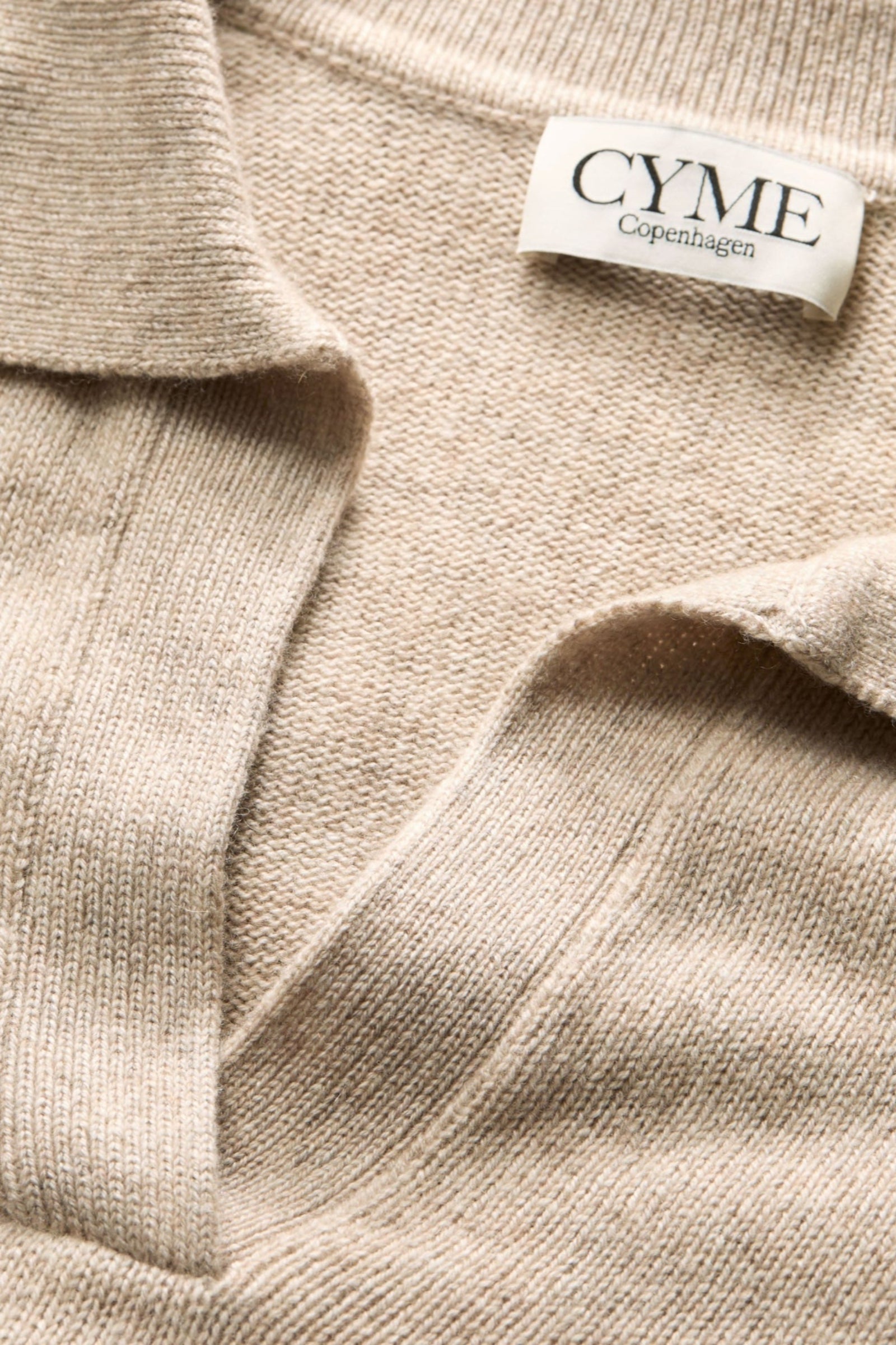 A close-up of the collar and neckline of a beige polo-style knit jumper, featuring a "CYME Copenhagen" label.
