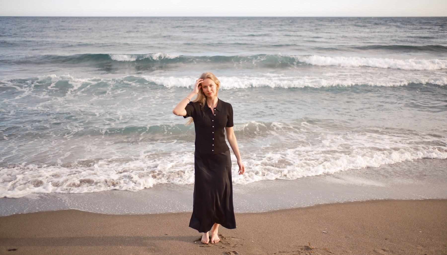 Woman wearing a black CYME pearl-button knit top and satin skirt on a quiet beach.