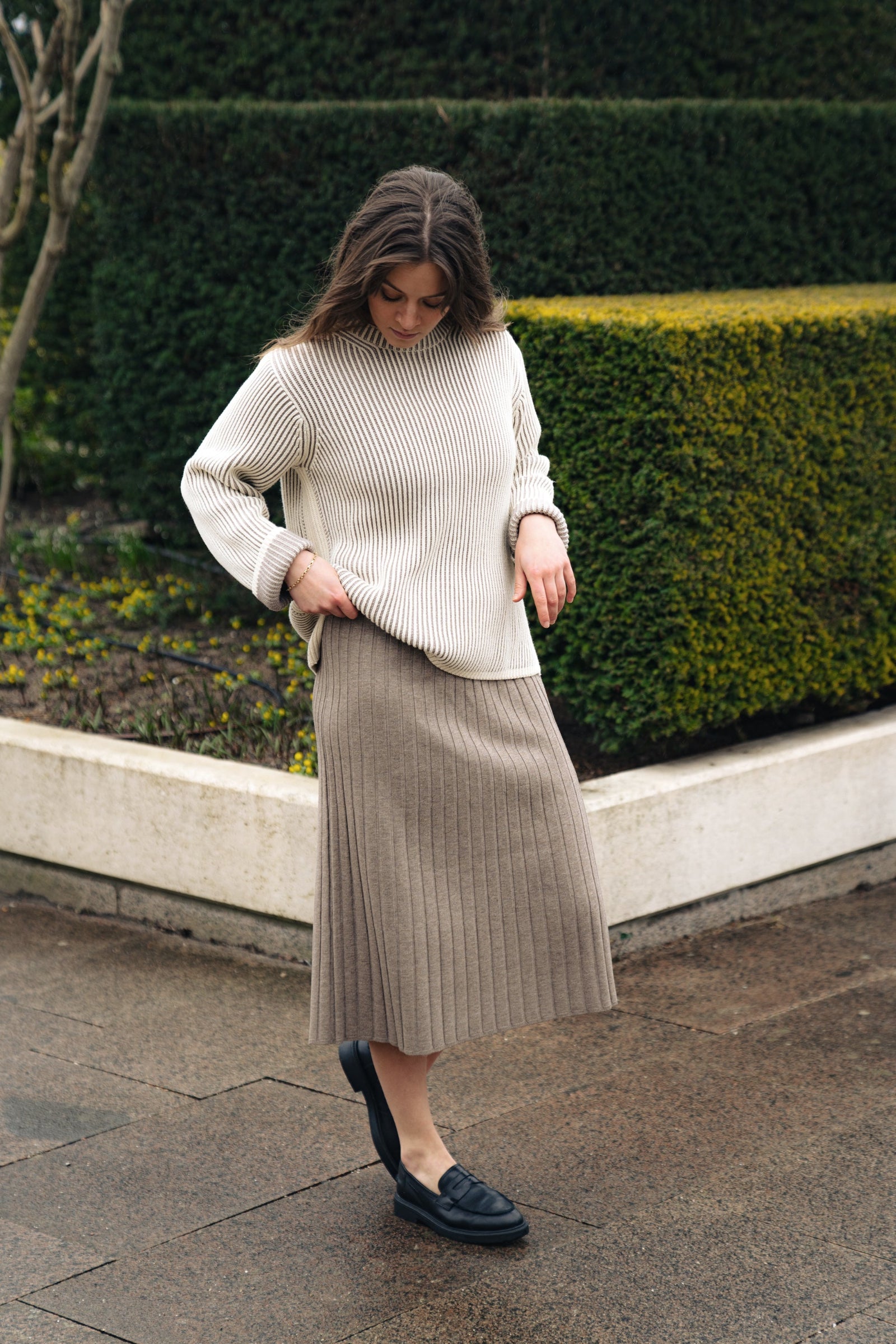 Woman wearing a cream sweater and beige pleated skirt standing outdoors.