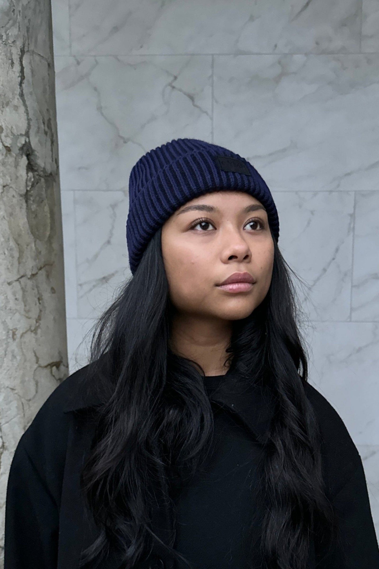 Woman wearing a dark navy knit beanie, standing in front of a marble column with long, wavy hair framing her face.
