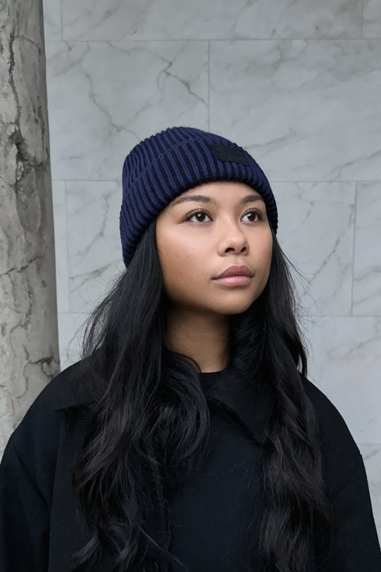 Woman wearing a dark navy knit beanie, standing in front of a marble column with long, wavy hair framing her face.