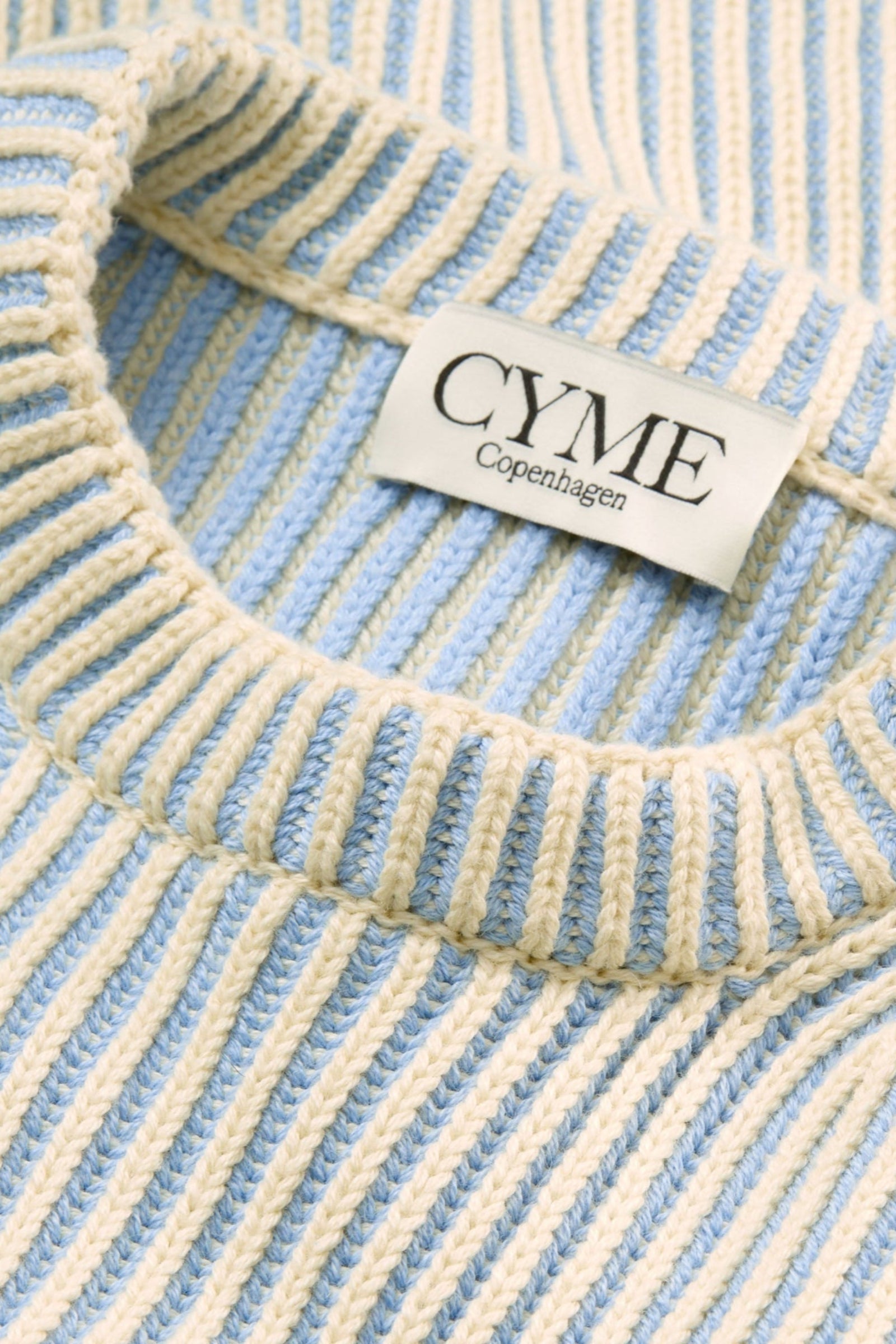 A close-up of the neckline and ribbed texture of a cream and light blue sweater, with a visible "CYME Copenhagen" label.