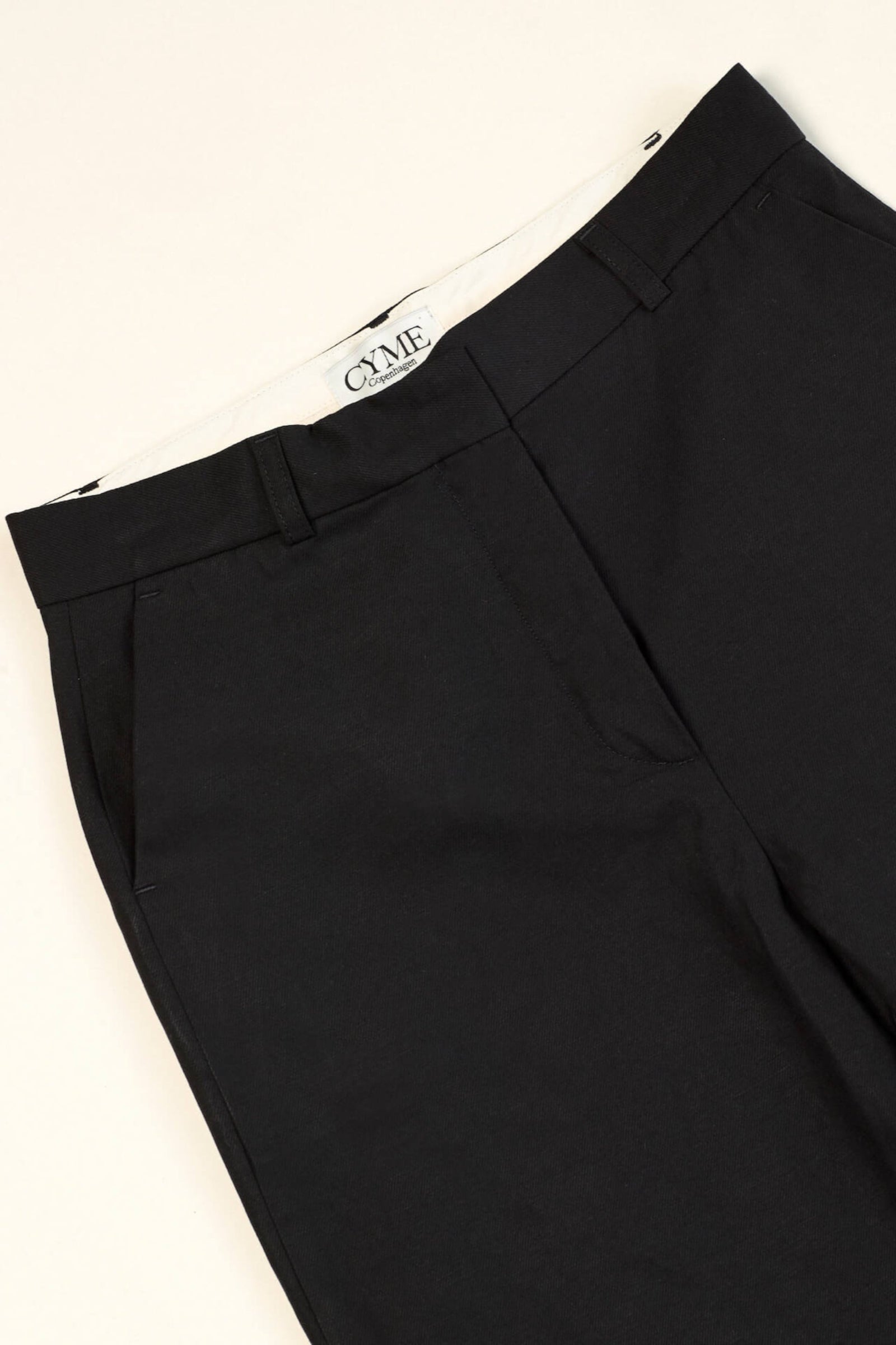Close-up of a pair of black trousers from CYME Copenhagen. The trousers feature a sleek, minimalistic design with a high waist and side pockets. The CYME Copenhagen label is prominently displayed on the inner waistband.