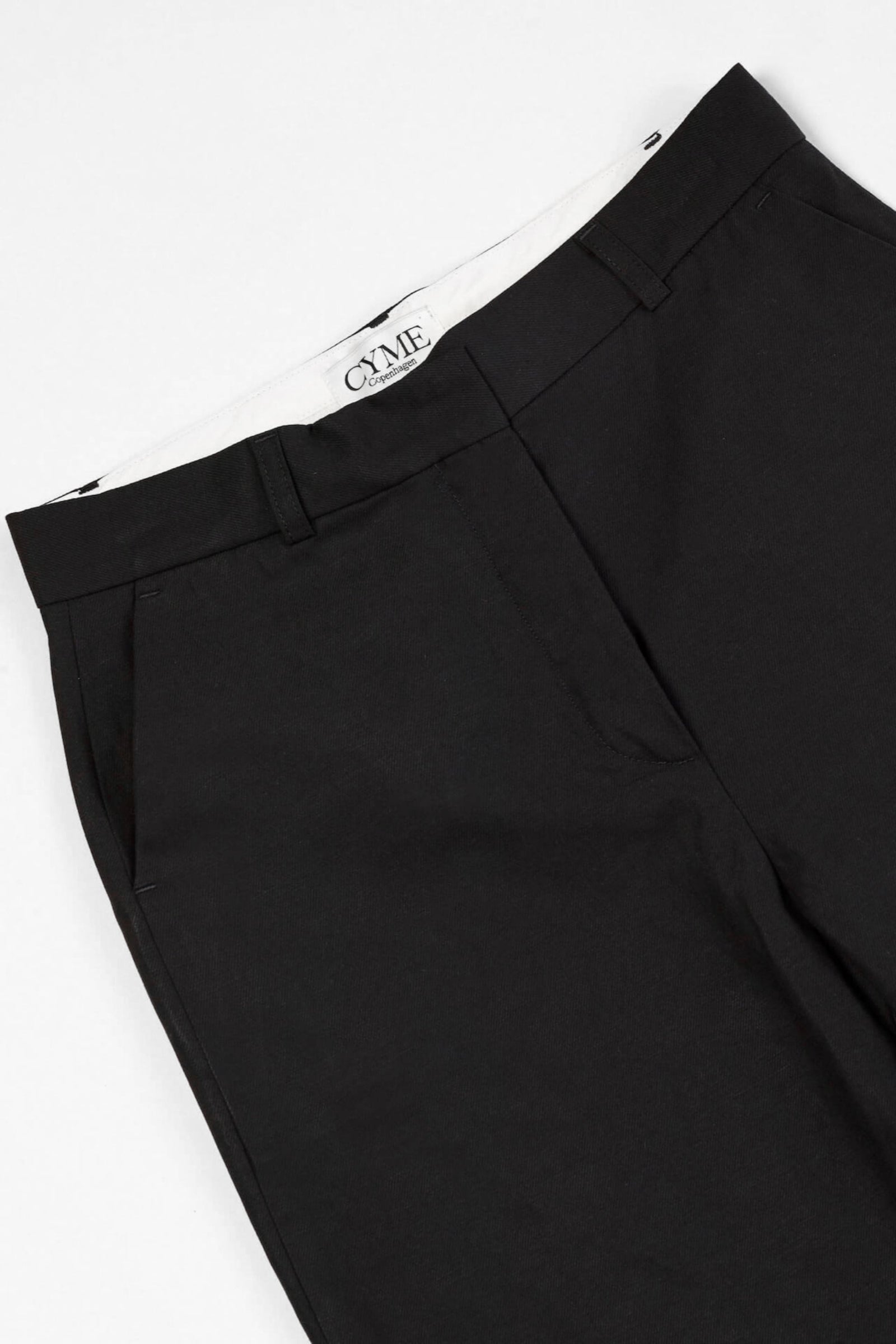 Close-up of a pair of black trousers from CYME Copenhagen. The trousers feature a sleek, minimalistic design with a high waist and side pockets. The CYME Copenhagen label is prominently displayed on the inner waistband.