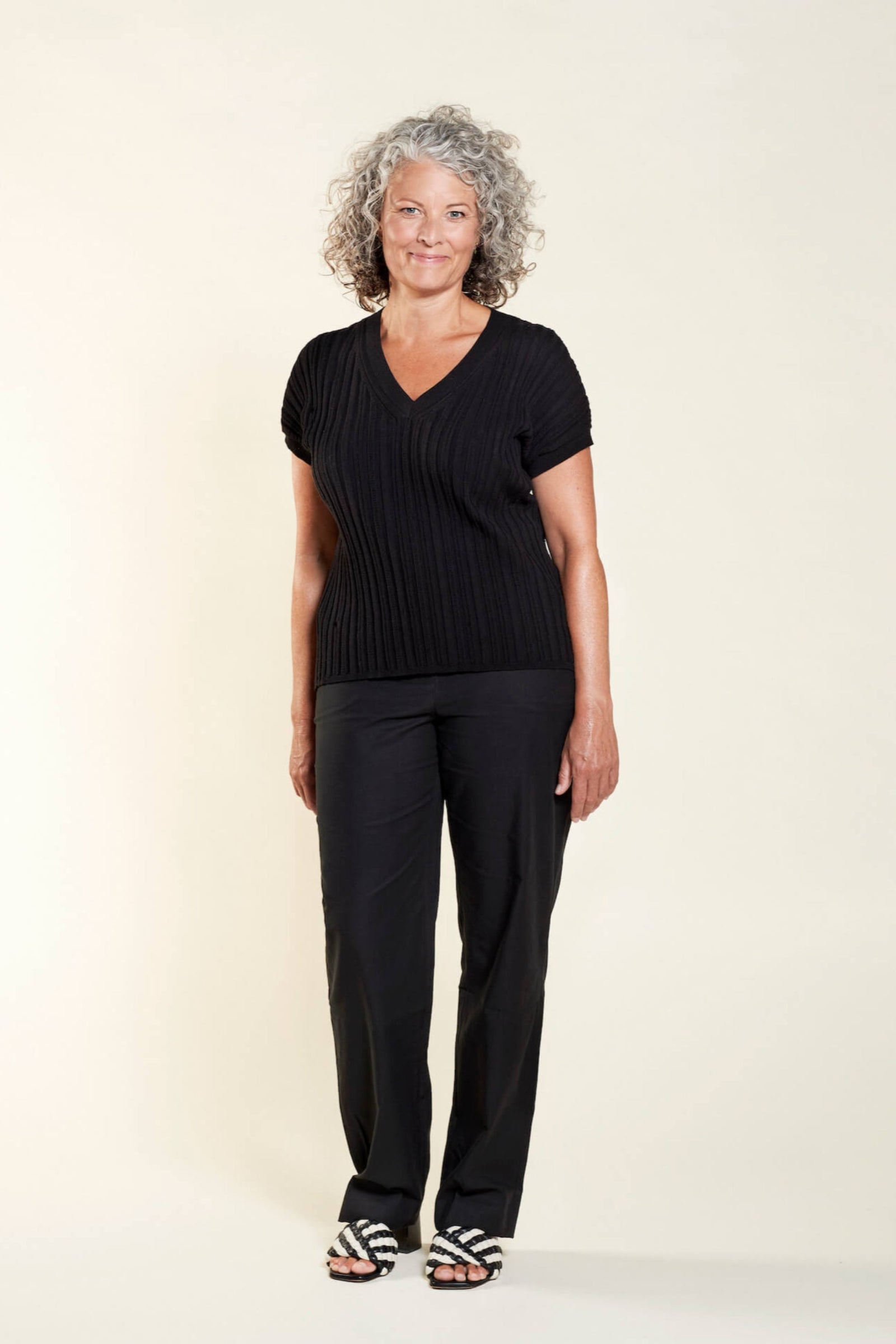 A woman with curly gray hair models a classic outfit from CYME Copenhagen. She is wearing a black V-neck short-sleeve sweater paired with black trousers. The look is completed with black and white striped sandals, emphasizing the brand's focus on timeless, high-quality fashion for all ages.