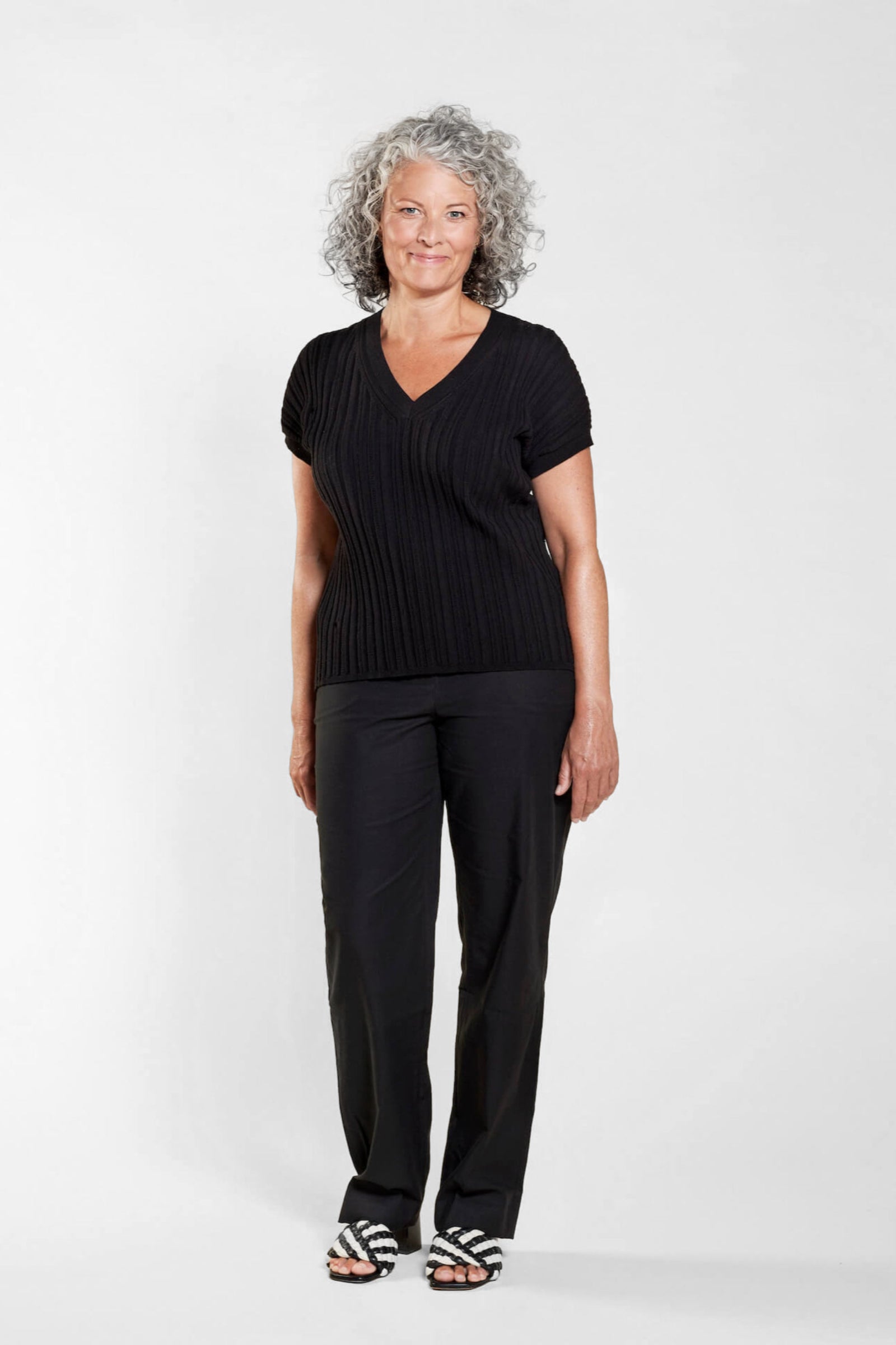 A woman with curly gray hair models a classic outfit from CYME Copenhagen. She is wearing a black V-neck short-sleeve sweater paired with black trousers. The look is completed with black and white striped sandals, emphasizing the brand's focus on timeless, high-quality fashion for all ages.