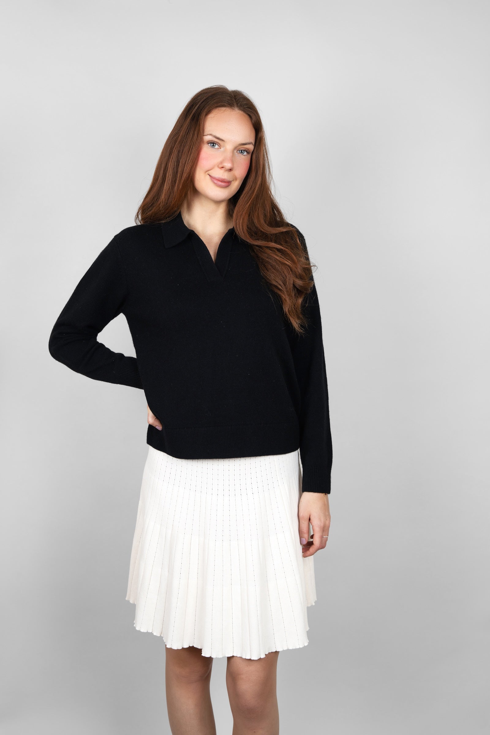 Front view of the CYME Cashmere-Merino Polo Jumper in black, styled with an ivory pleated skirt. This relaxed polo jumper is knitted in a warm, soft cashmere-merino blend.