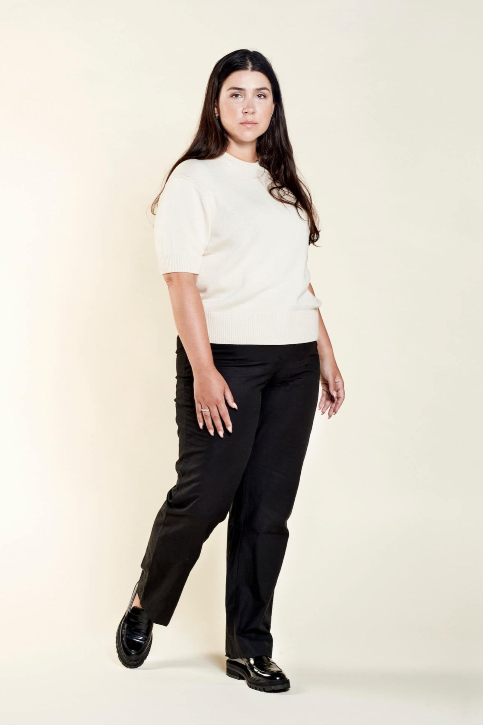 A woman with long dark hair models a casual outfit from CYME Copenhagen. She is wearing a cream-colored short-sleeve sweater paired with black trousers. The look is completed with black loafers, emphasizing the brand's focus on timeless, high-quality fashion.