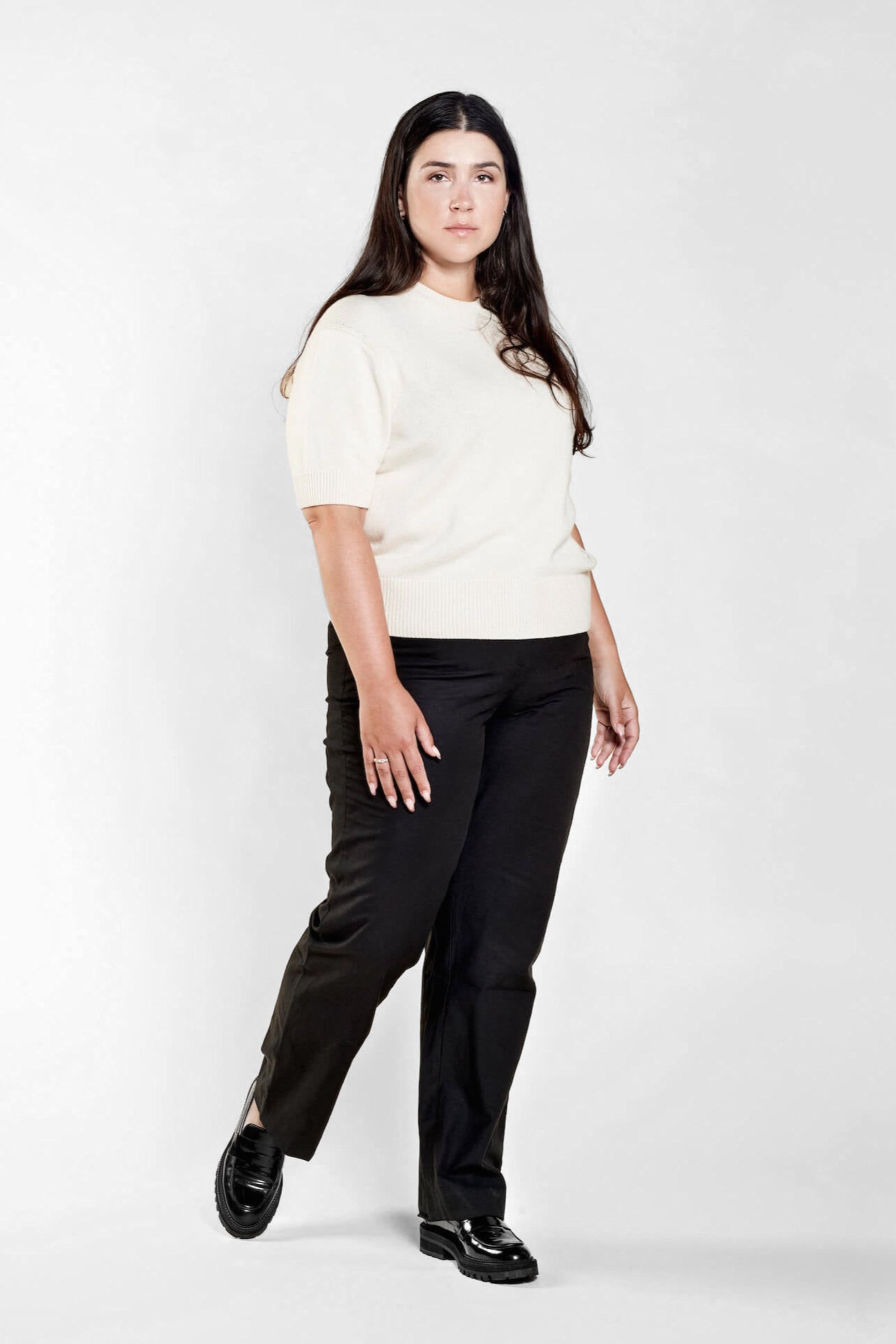 A woman with long dark hair models a casual outfit from CYME Copenhagen. She is wearing a cream-colored short-sleeve sweater paired with black trousers. The look is completed with black loafers, emphasizing the brand's focus on timeless, high-quality fashion.