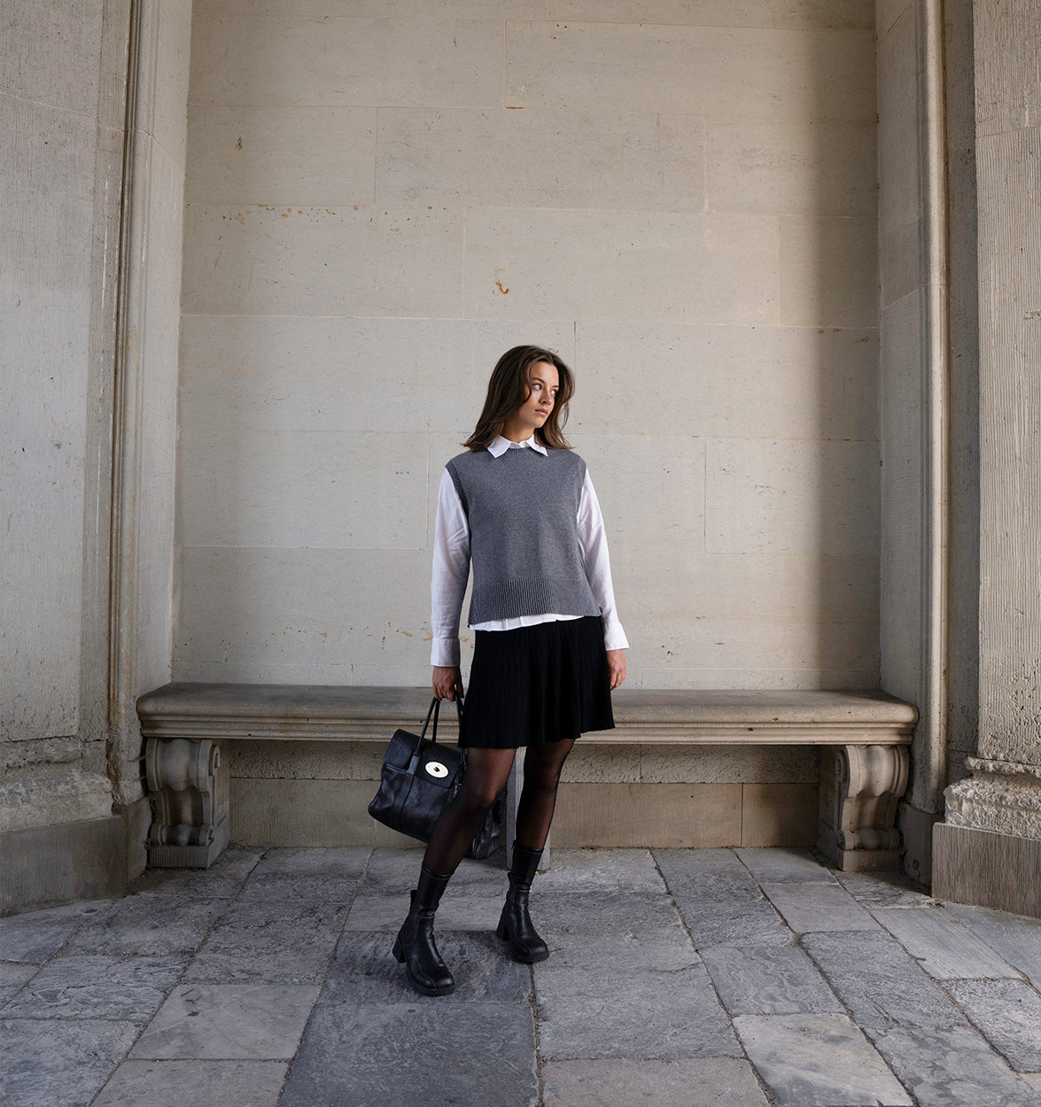 Woman wearing a grey vest and black short skirt against a beige wall.