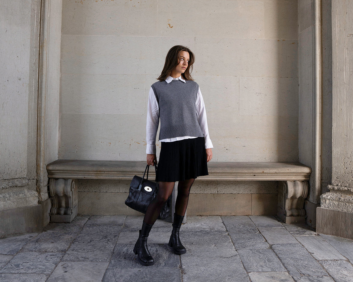 Woman wearing a grey vest and black short skirt against a beige wall.