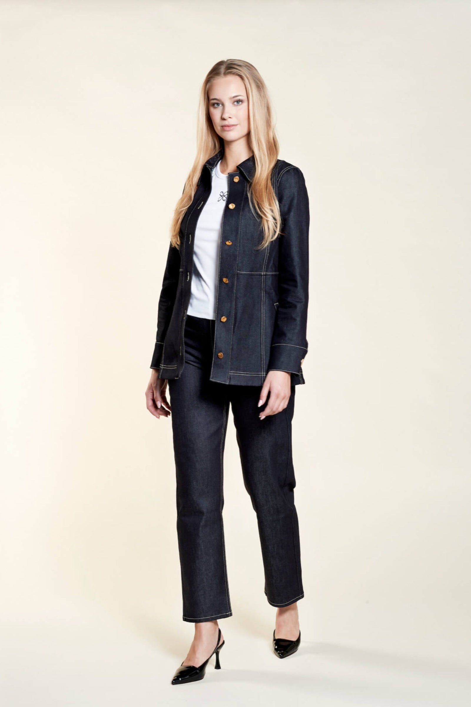 A woman with long blonde hair models a dark denim jacket and matching jeans from CYME Copenhagen. She wears a white top underneath, showcasing a subtle embroidered logo. The outfit is complemented by black heels, emphasizing the brand's focus on timeless, high-quality fashion.