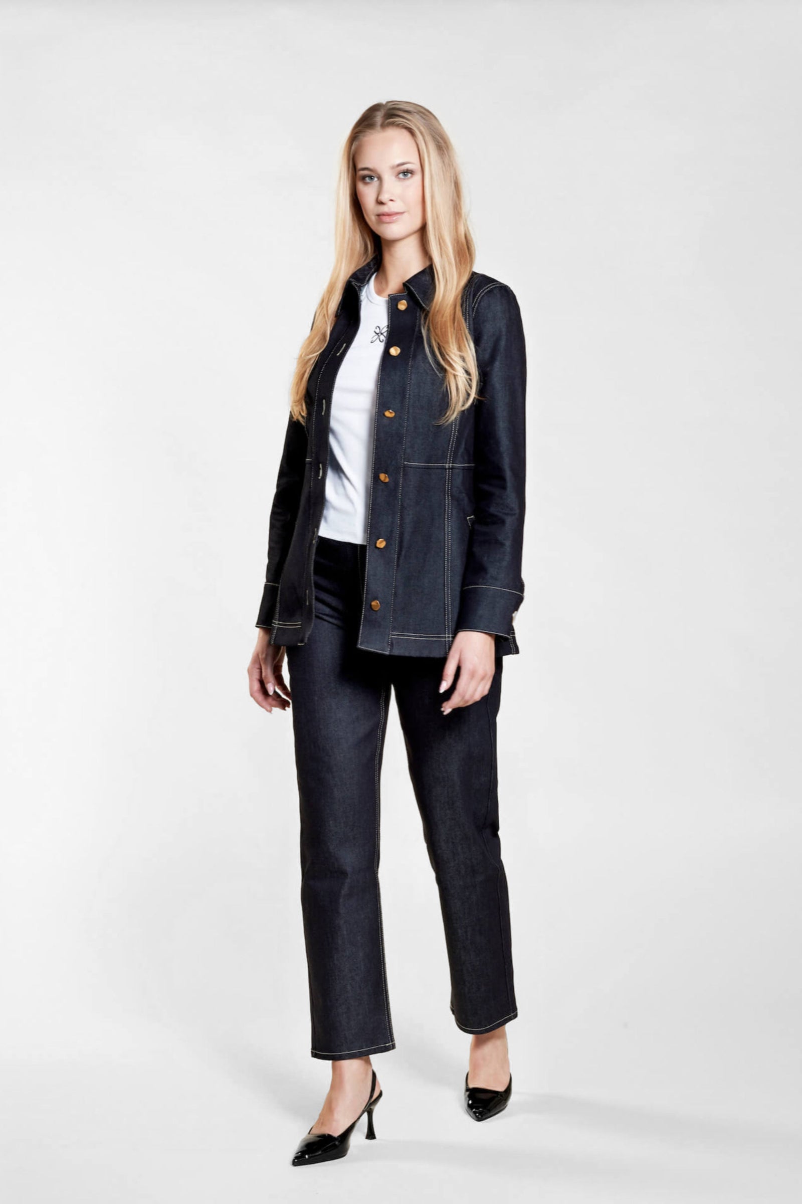 A woman with long blonde hair models a dark denim jacket and matching jeans from CYME Copenhagen. She wears a white top underneath, showcasing a subtle embroidered logo. The outfit is complemented by black heels, emphasizing the brand's focus on timeless, high-quality fashion.