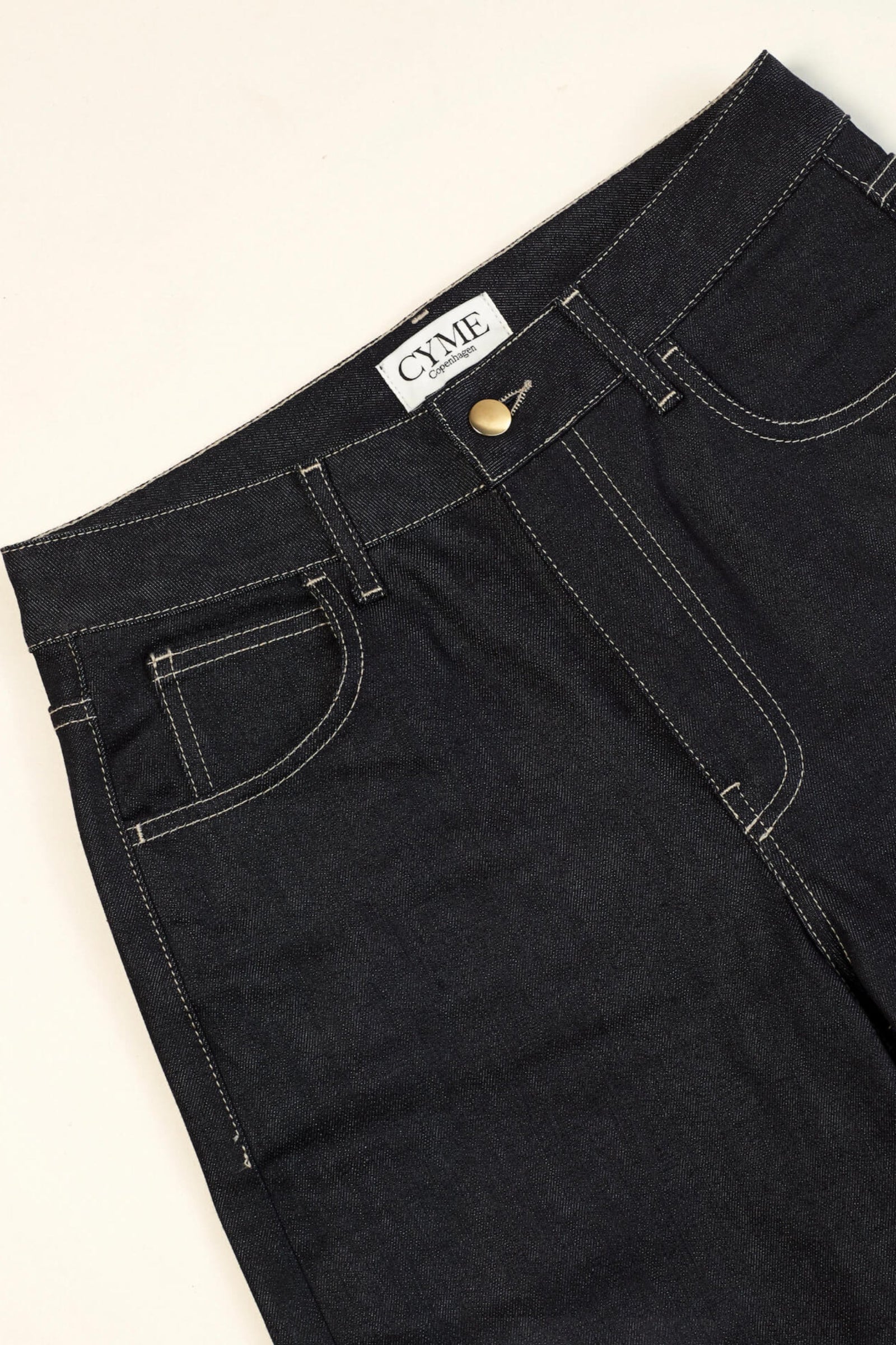Close-up of a pair of dark blue high-waisted jeans from CYME Copenhagen. The jeans feature a classic five-pocket design with contrasting white stitching and a brass button closure. The CYME Copenhagen label is prominently displayed on the inner waistband.