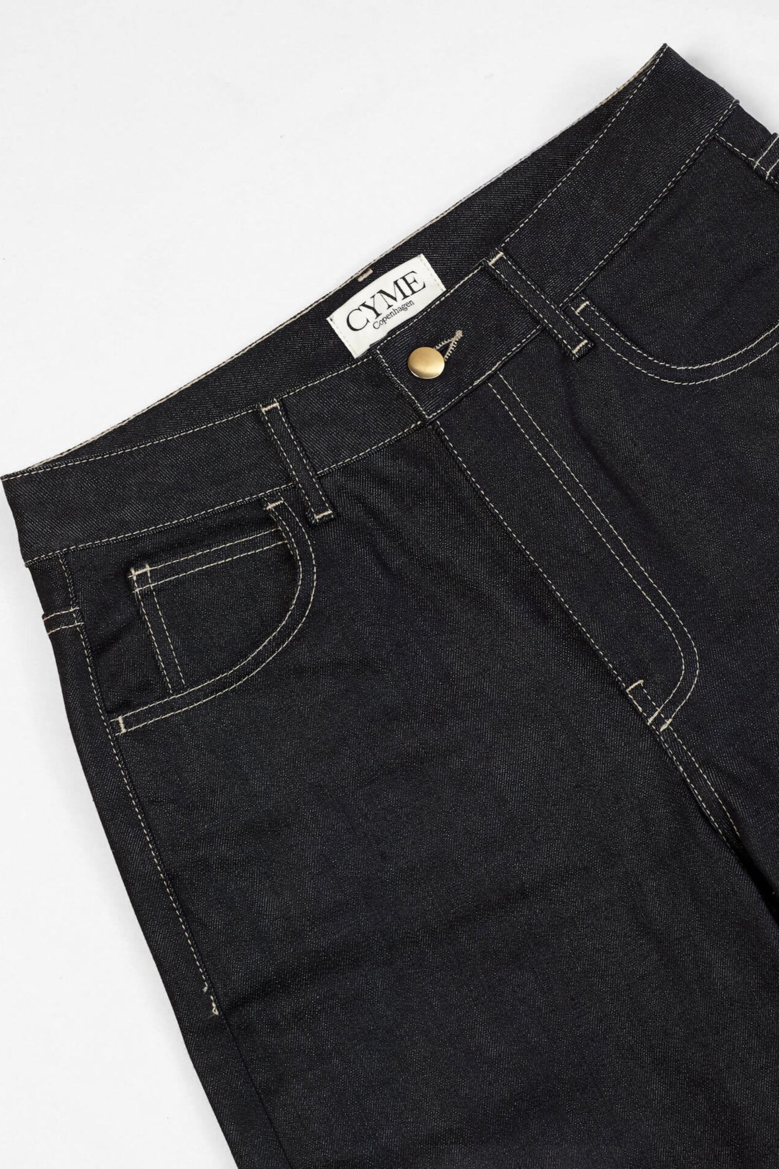 Close-up of a pair of dark blue high-waisted jeans from CYME Copenhagen. The jeans feature a classic five-pocket design with contrasting white stitching and a brass button closure. The CYME Copenhagen label is prominently displayed on the inner waistband.