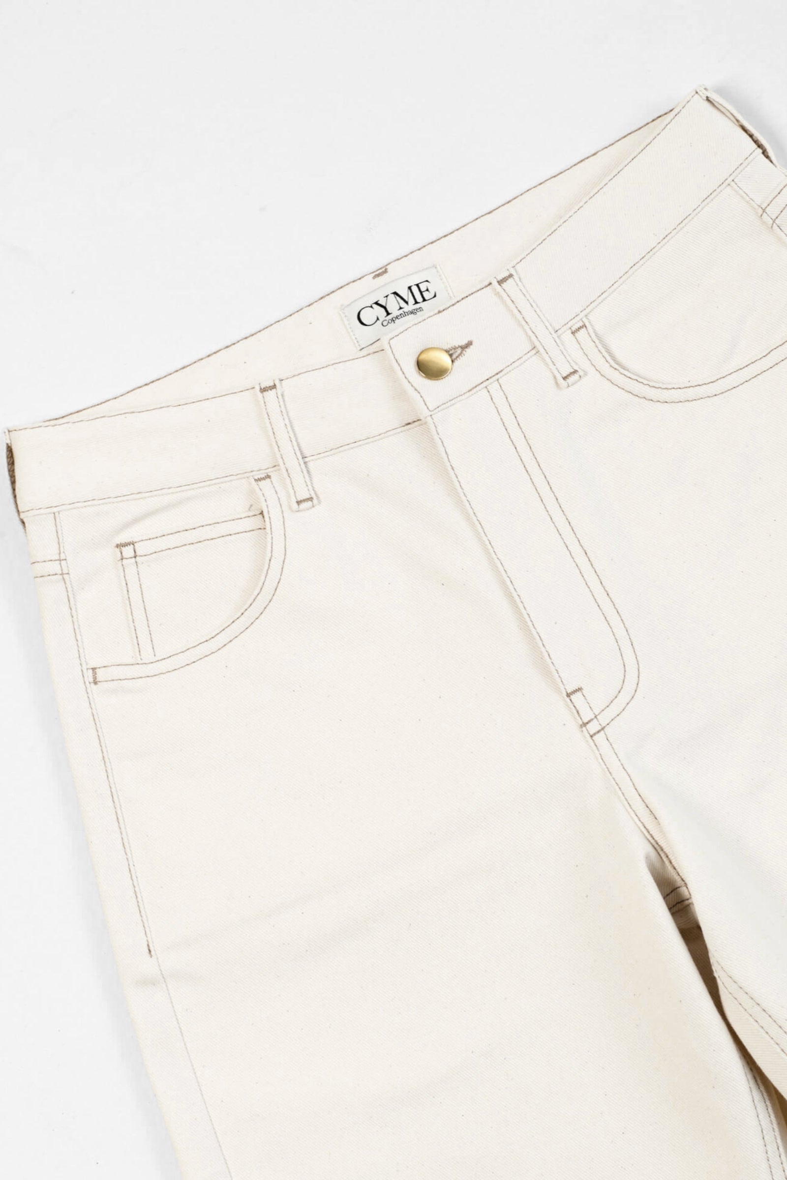 Close-up of a pair of beige high-waisted jeans from CYME Copenhagen. The jeans feature a classic five-pocket design, with visible stitching and a brass button closure. The CYME Copenhagen label is prominently displayed on the inner waistband.