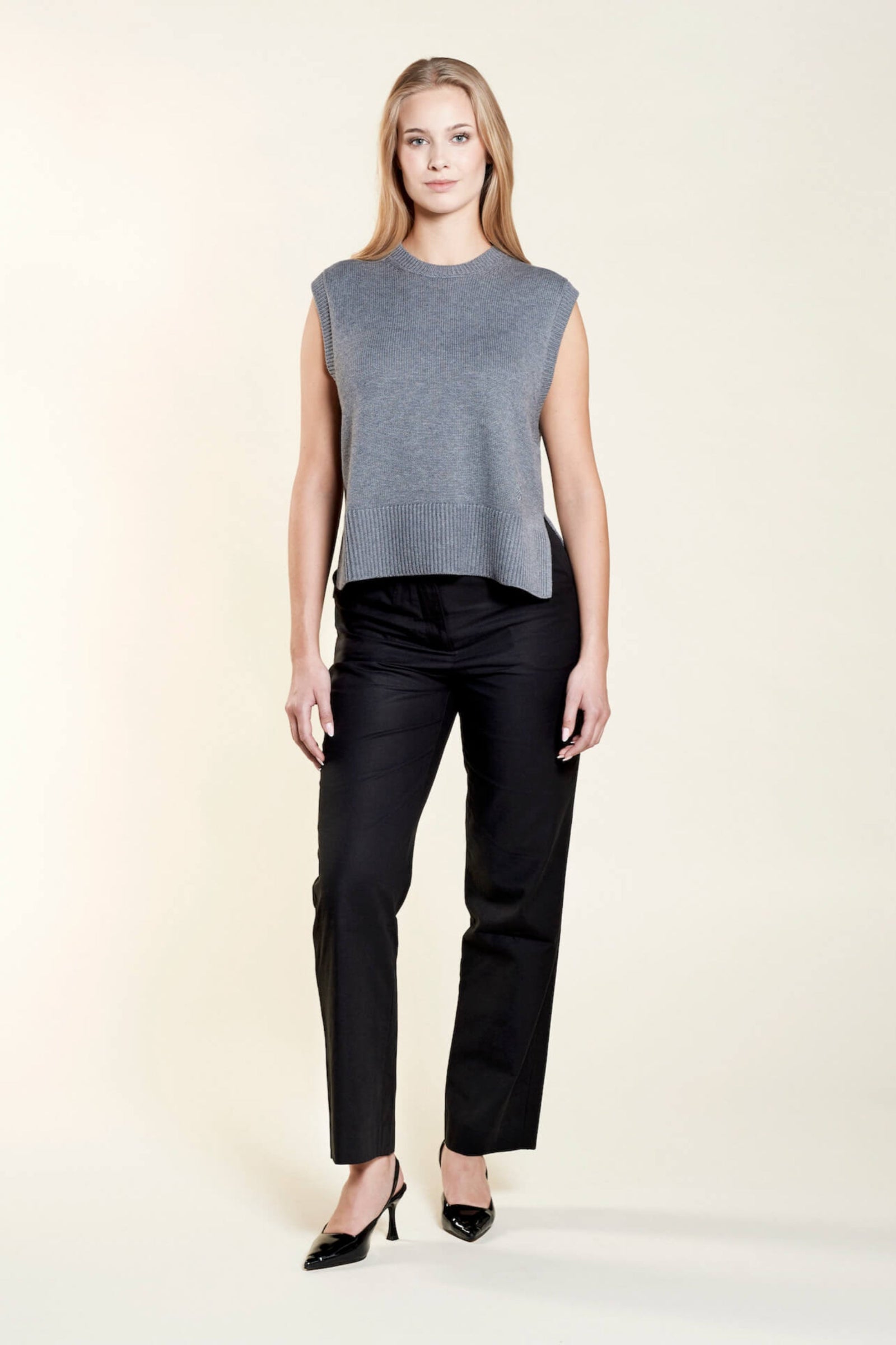 A woman with long blonde hair models a chic outfit from CYME Copenhagen. She is wearing a sleeveless gray knit top paired with black trousers and black heels, showcasing a minimalist and sophisticated look that emphasizes the brand's commitment to timeless fashion.