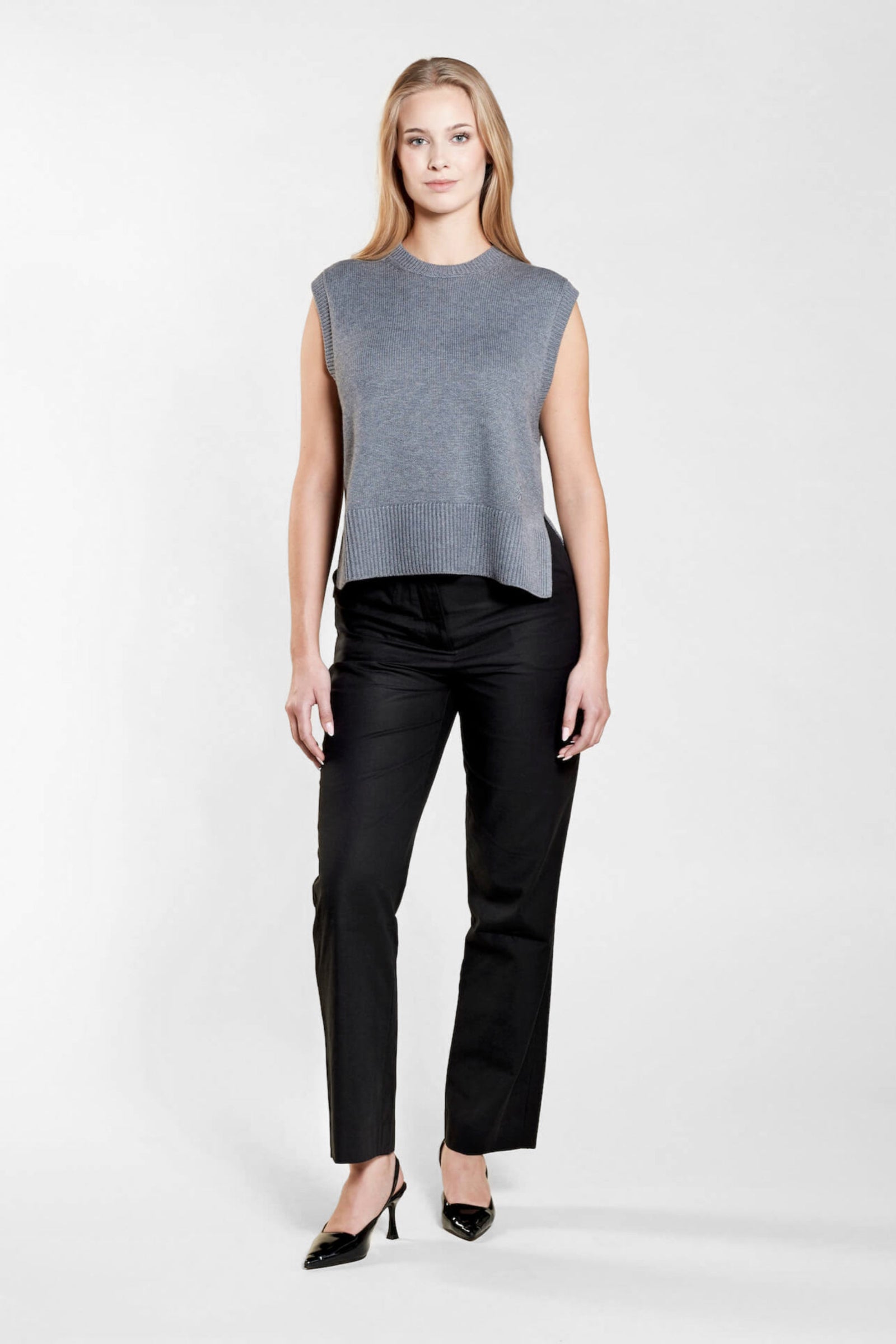 A woman with long blonde hair models a chic outfit from CYME Copenhagen. She is wearing a sleeveless gray knit top paired with black trousers and black heels, showcasing a minimalist and sophisticated look that emphasizes the brand's commitment to timeless fashion.