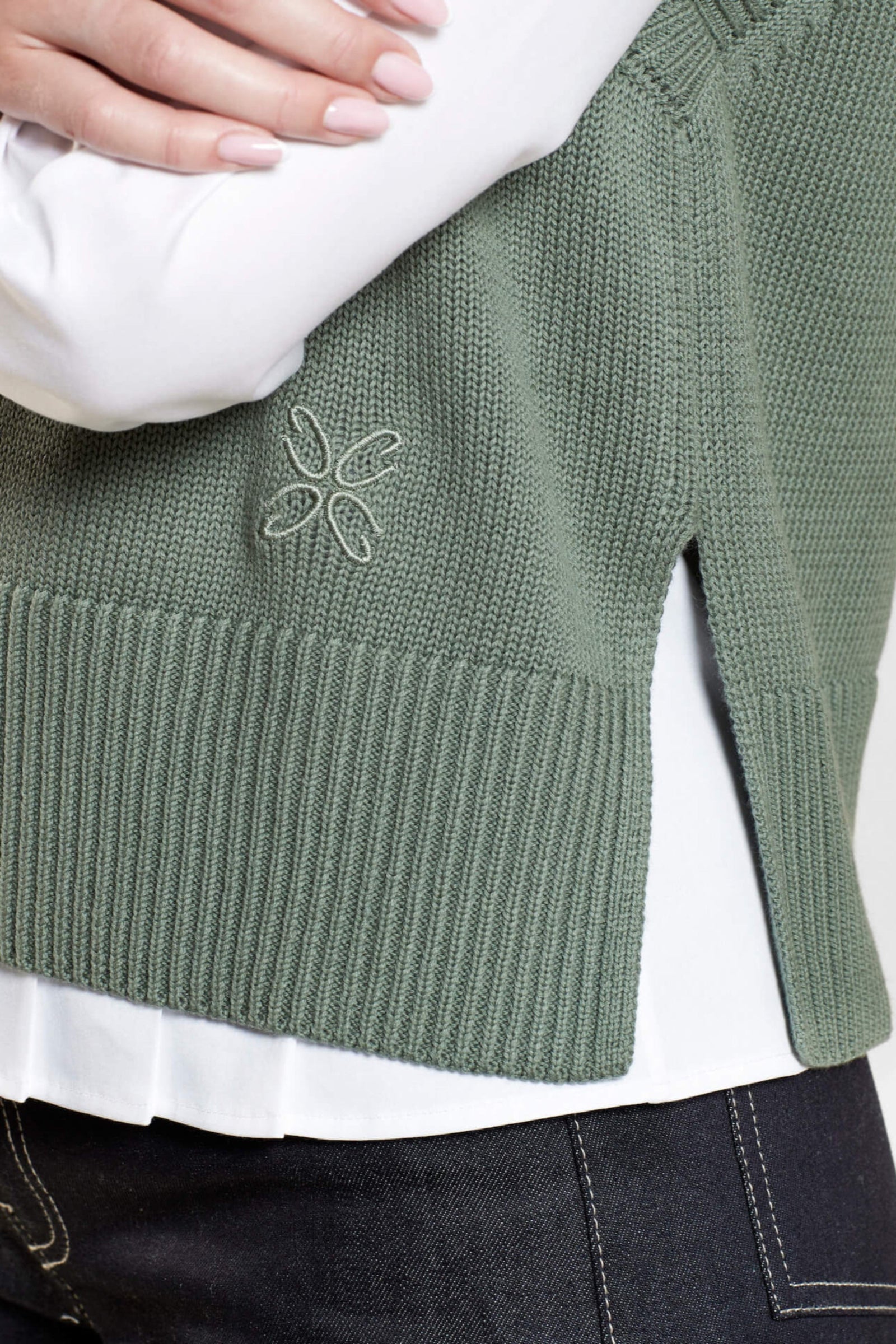 Close-up of a green knit vest from CYME Copenhagen, featuring a side slit and subtle embroidered logo on the lower side, layered over a white shirt and dark denim jeans. The image highlights the intricate details and quality of the garment.