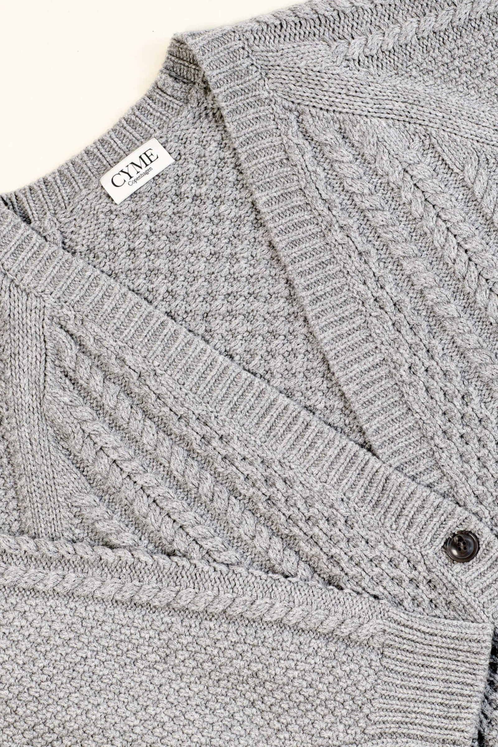 Close-up of a light grey knitted cardigan from CYME Copenhagen. The cardigan features intricate cable knit and textured patterns with a V-neckline and black buttons. The CYME Copenhagen label is prominently displayed on the inner neckline.