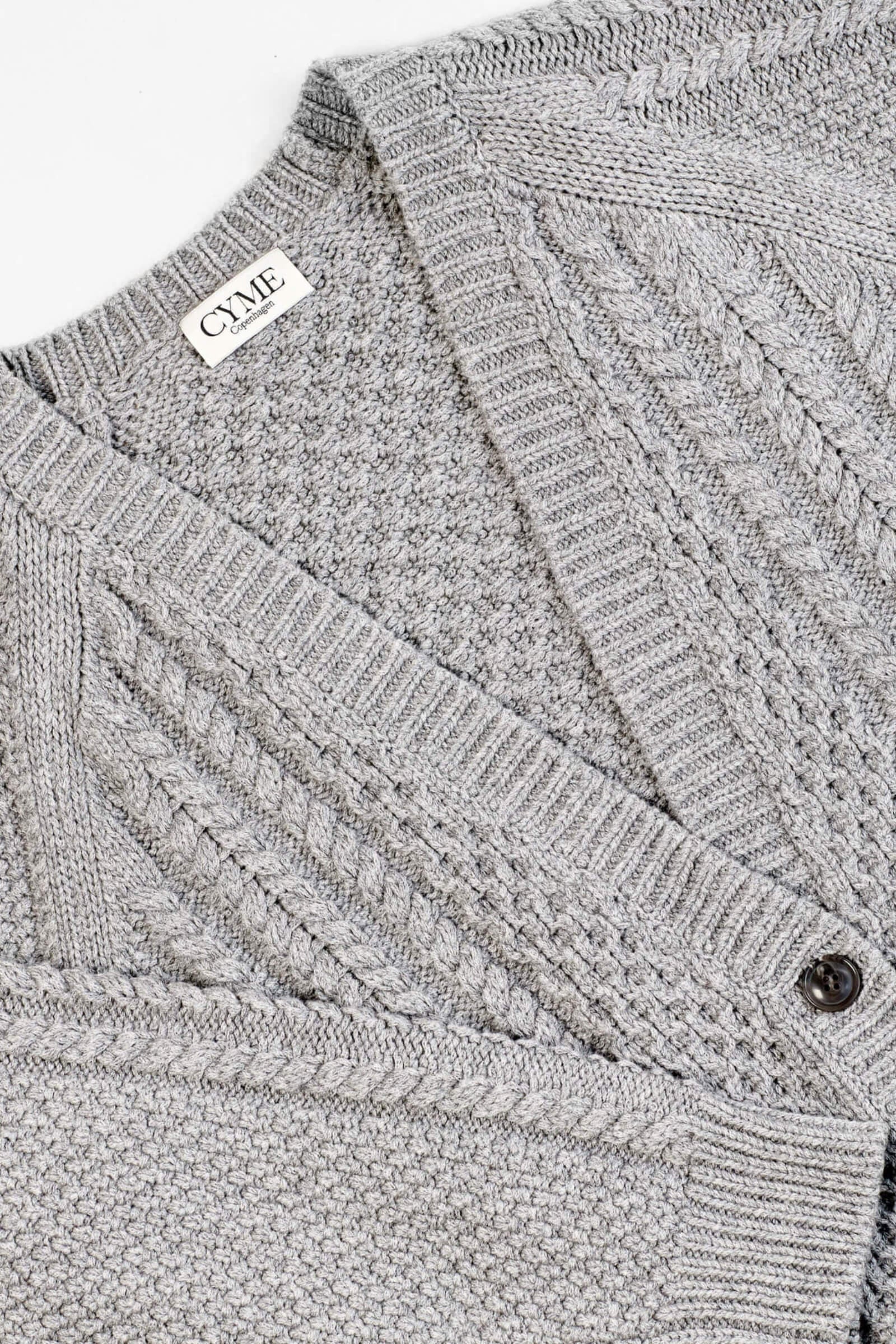 Close-up of a light grey knitted cardigan from CYME Copenhagen. The cardigan features intricate cable knit and textured patterns with a V-neckline and black buttons. The CYME Copenhagen label is prominently displayed on the inner neckline.