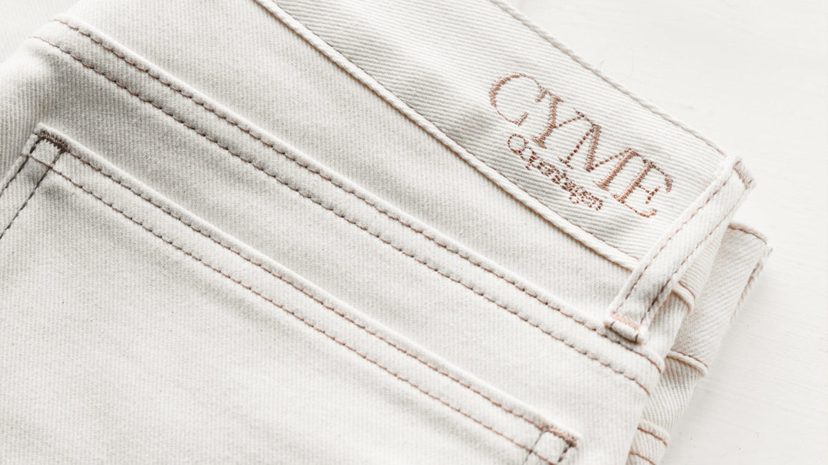 White jeans with 'CYME Copenhagen' branding on a beige background