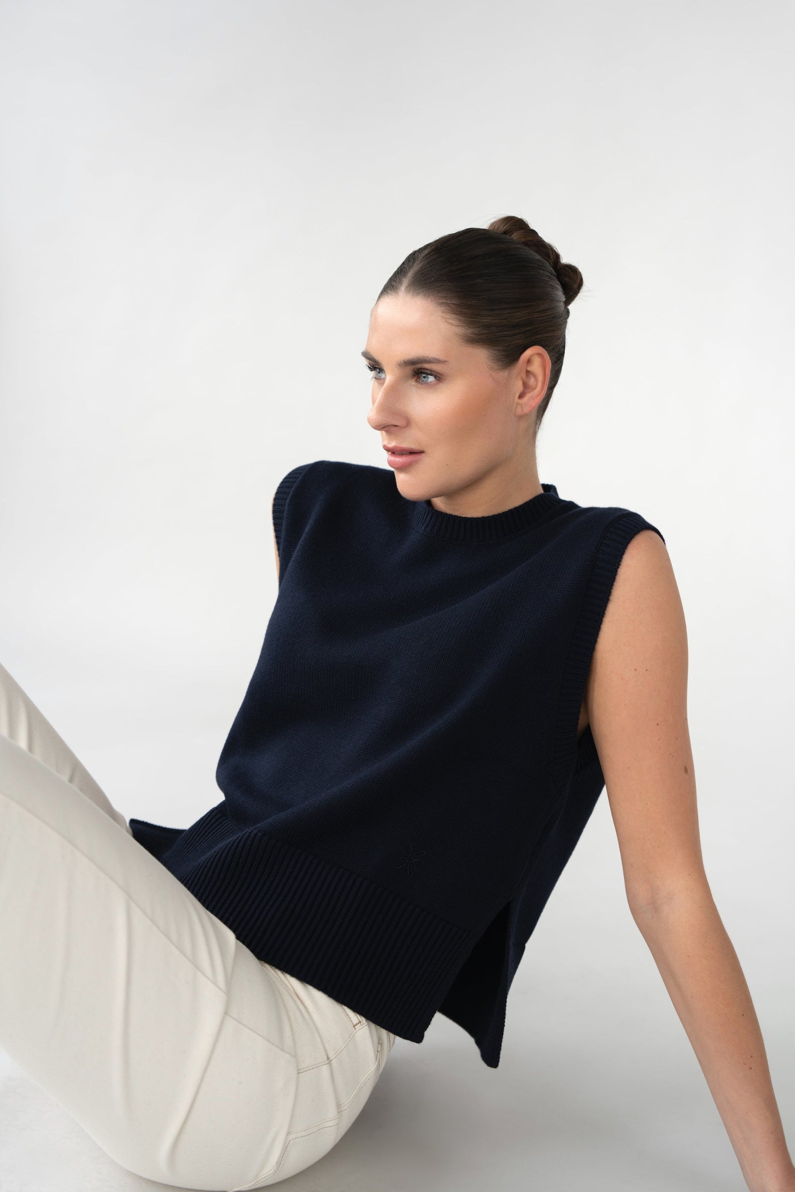 On-model seated view of women's sleeveless knitted vest, navy, crew neck and wide hem, studio.