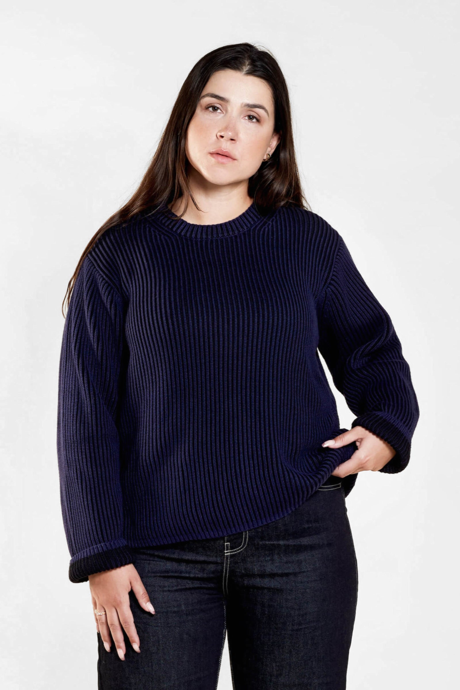 Portrait of a woman with long dark hair wearing a navy blue ribbed sweater and dark jeans. She is standing against a light background, looking directly at the camera with one hand resting on her hip.