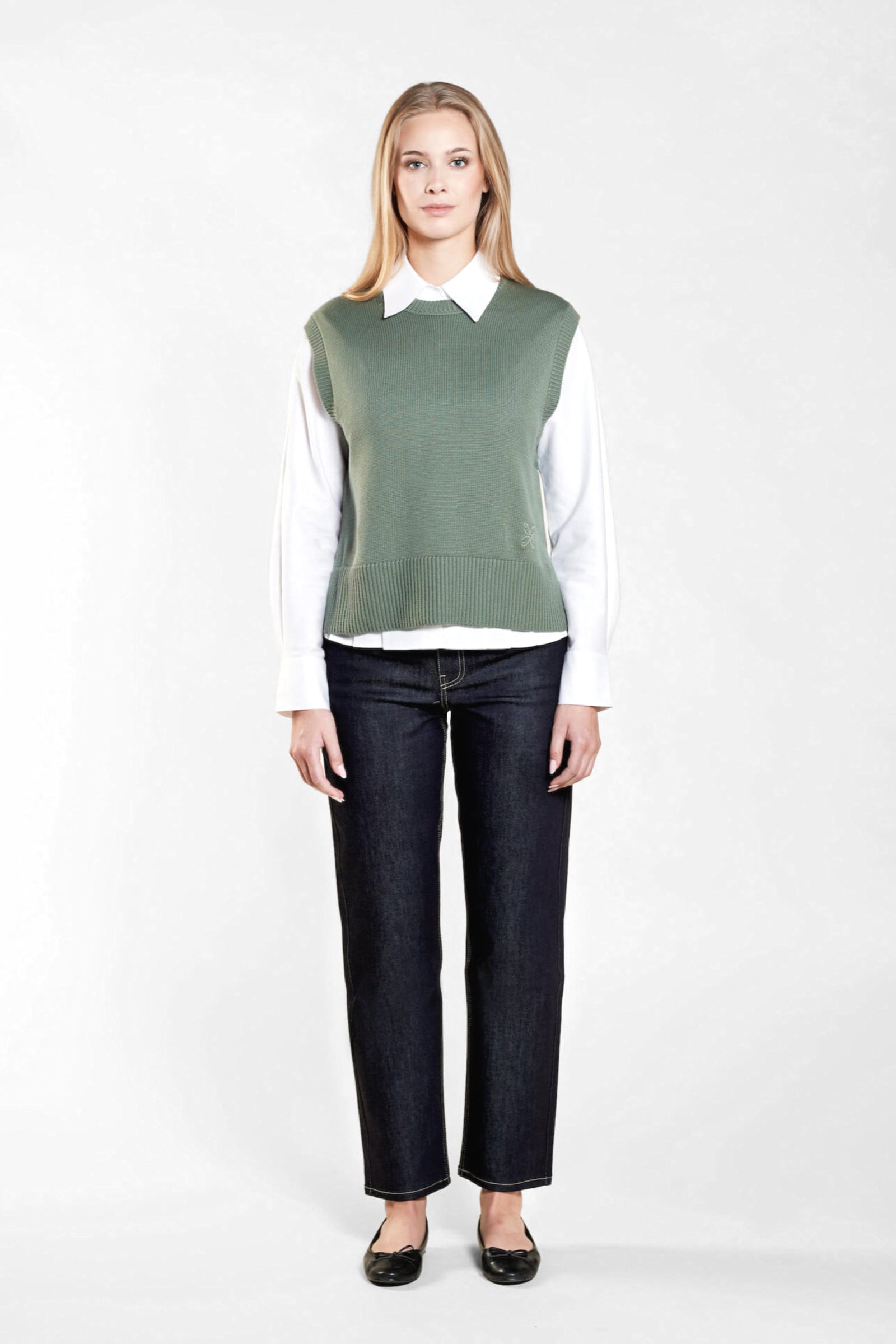 A woman with long blonde hair models a stylish outfit from CYME Copenhagen. She is wearing a sage green sleeveless knit vest over a white collared shirt, paired with dark blue jeans and black ballet flats. The outfit highlights the brand's focus on timeless and versatile fashion.