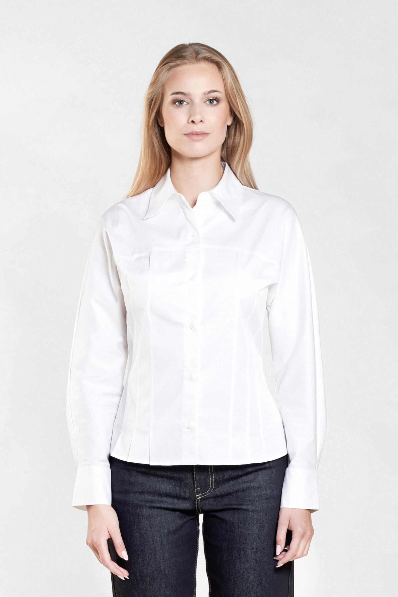 Front view of a woman with long blonde hair modeling a white button-down shirt paired with dark blue jeans from CYME Copenhagen. The image highlights the shirt's tailored fit and classic design, emphasizing the brand's commitment to high-quality, timeless fashion.