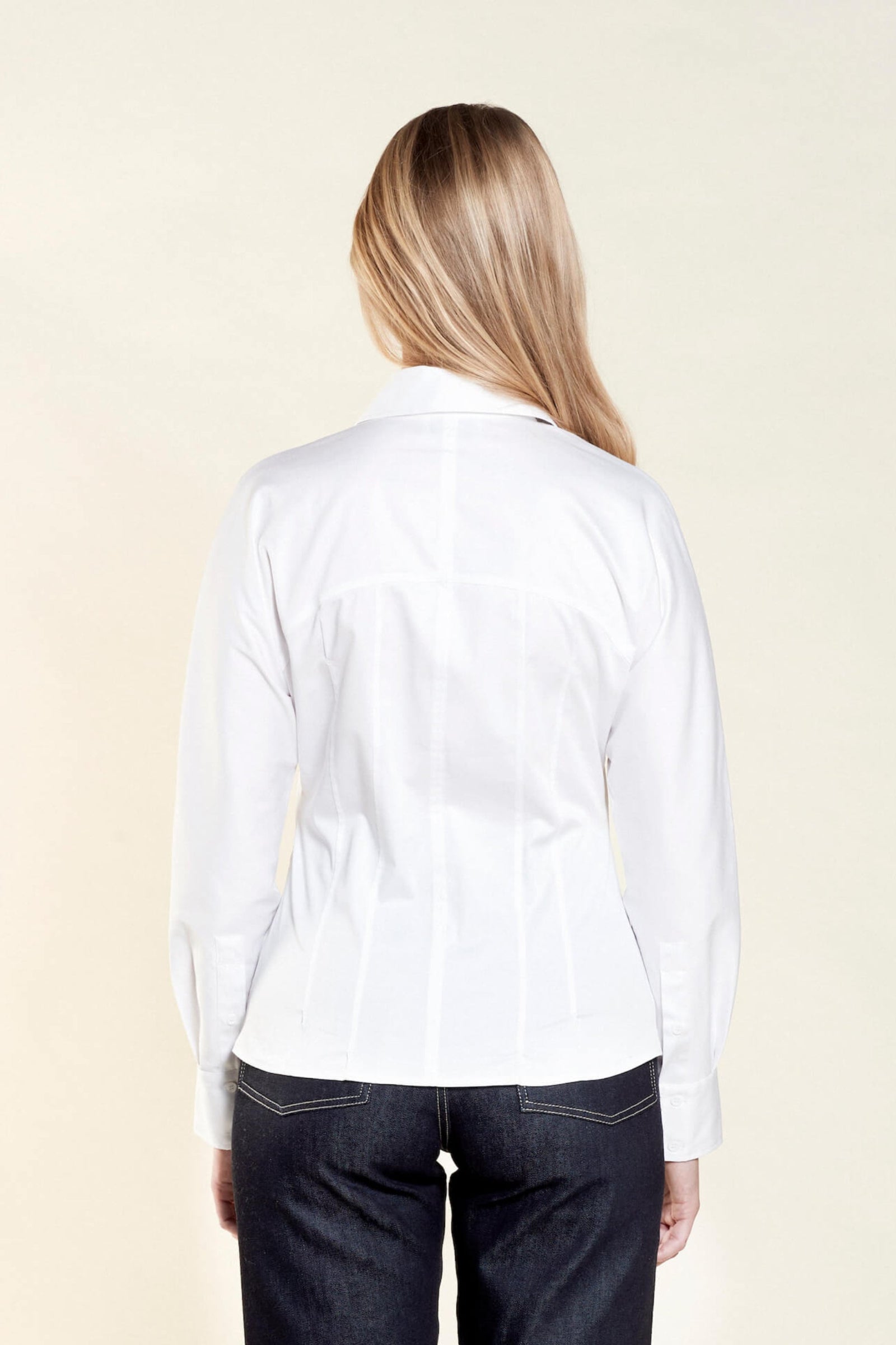 Rear view of a woman with long blonde hair modeling a white button-down shirt paired with dark blue jeans from CYME Copenhagen. The image highlights the shirt's tailored fit and clean lines, emphasizing the brand's focus on high-quality craftsmanship.