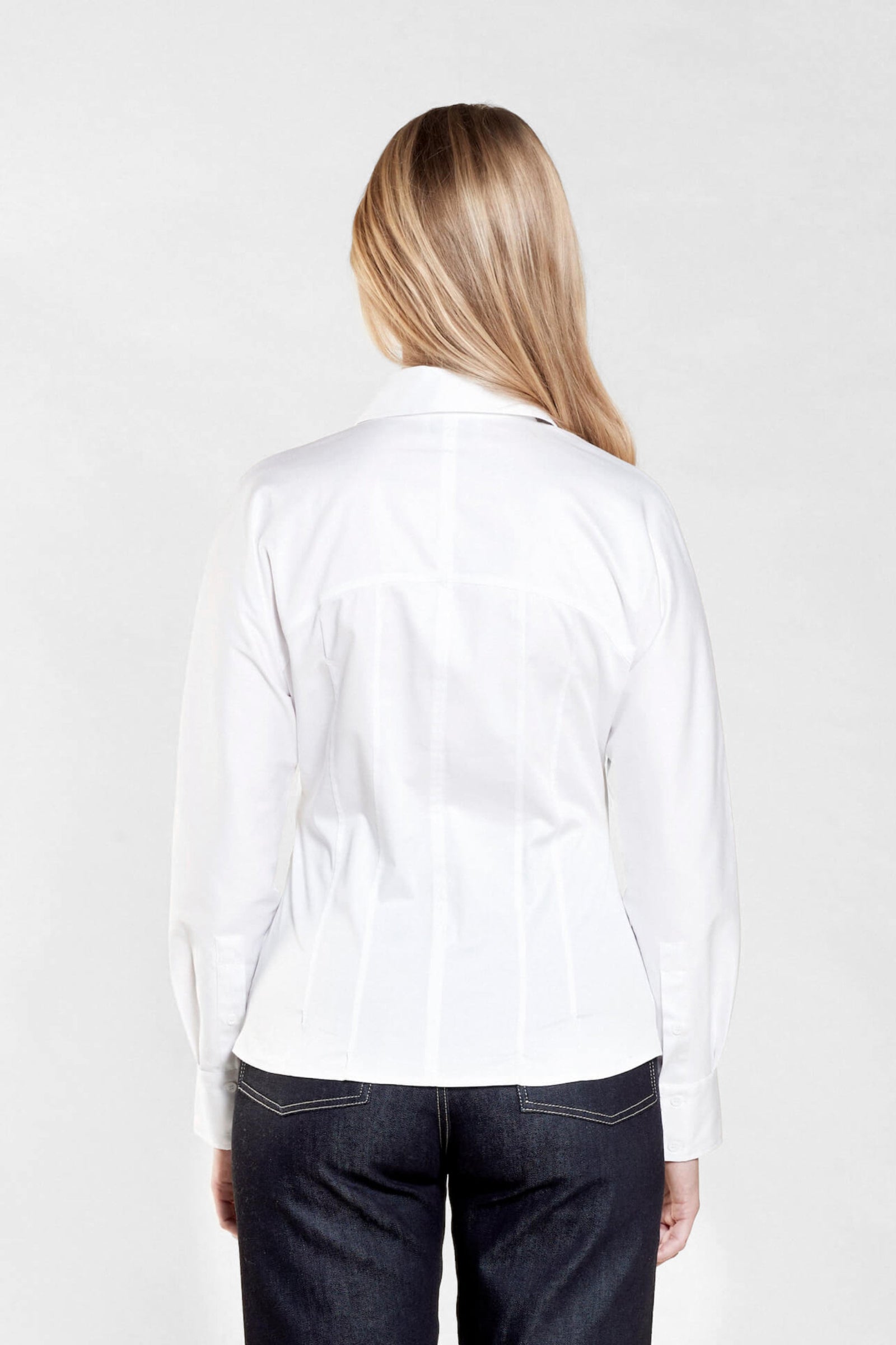 Rear view of a woman with long blonde hair modeling a white button-down shirt paired with dark blue jeans from CYME Copenhagen. The image highlights the shirt's tailored fit and clean lines, emphasizing the brand's focus on high-quality craftsmanship.