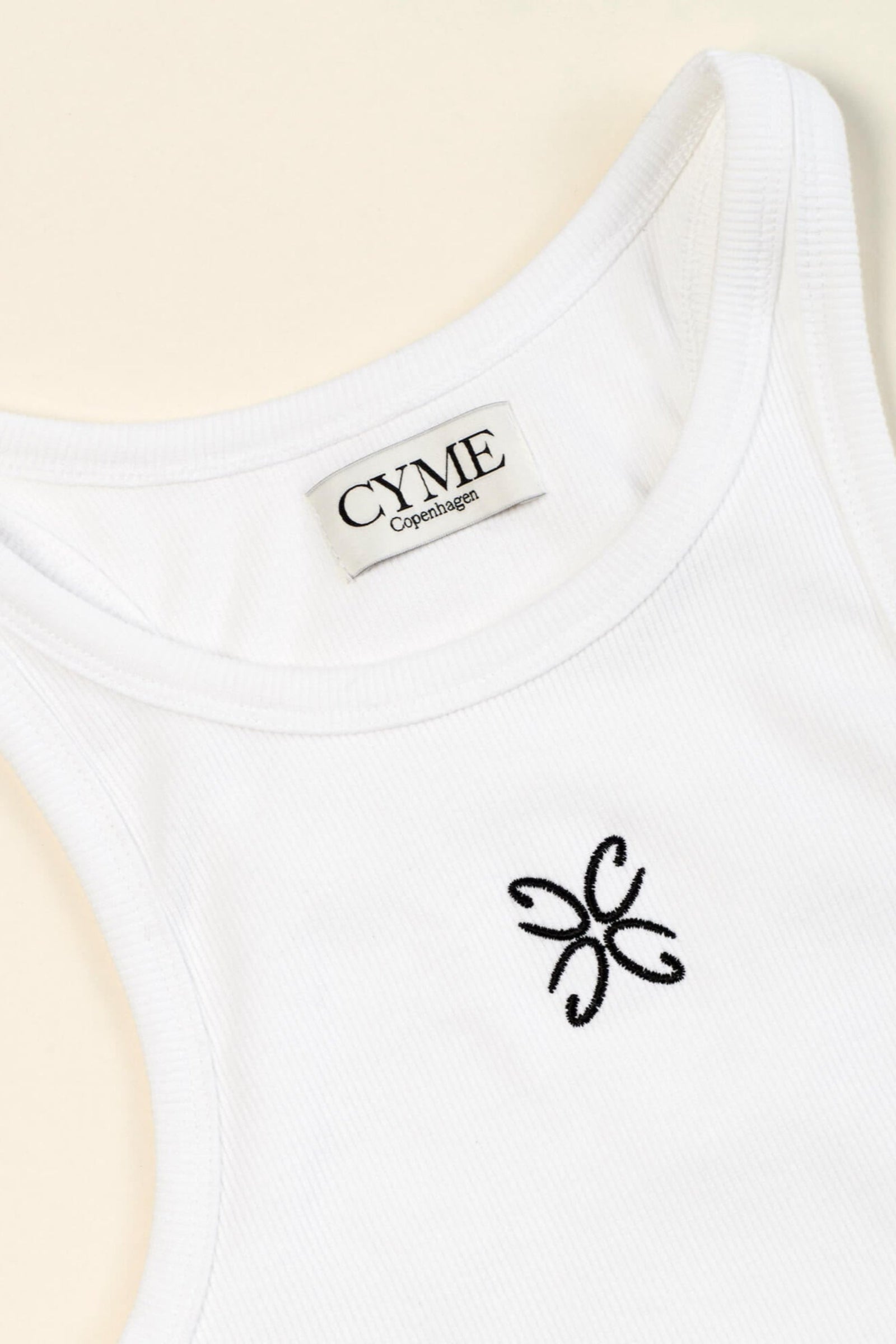 Close-up view of a white tank top with the CYME Copenhagen label sewn on the inside of the neckline and a delicate black embroidered logo on the chest. The tank top features a ribbed texture, highlighting the brand's focus on high-quality materials and timeless design.