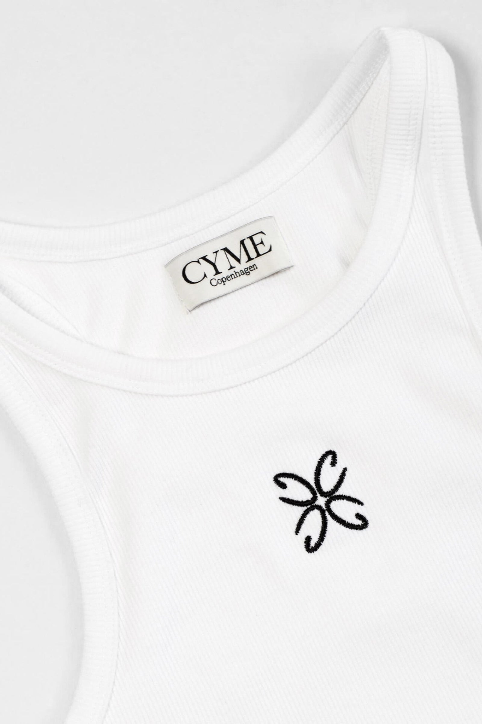 Close-up view of a white tank top with the CYME Copenhagen label sewn on the inside of the neckline and a delicate black embroidered logo on the chest. The tank top features a ribbed texture, highlighting the brand's focus on high-quality materials and timeless design.