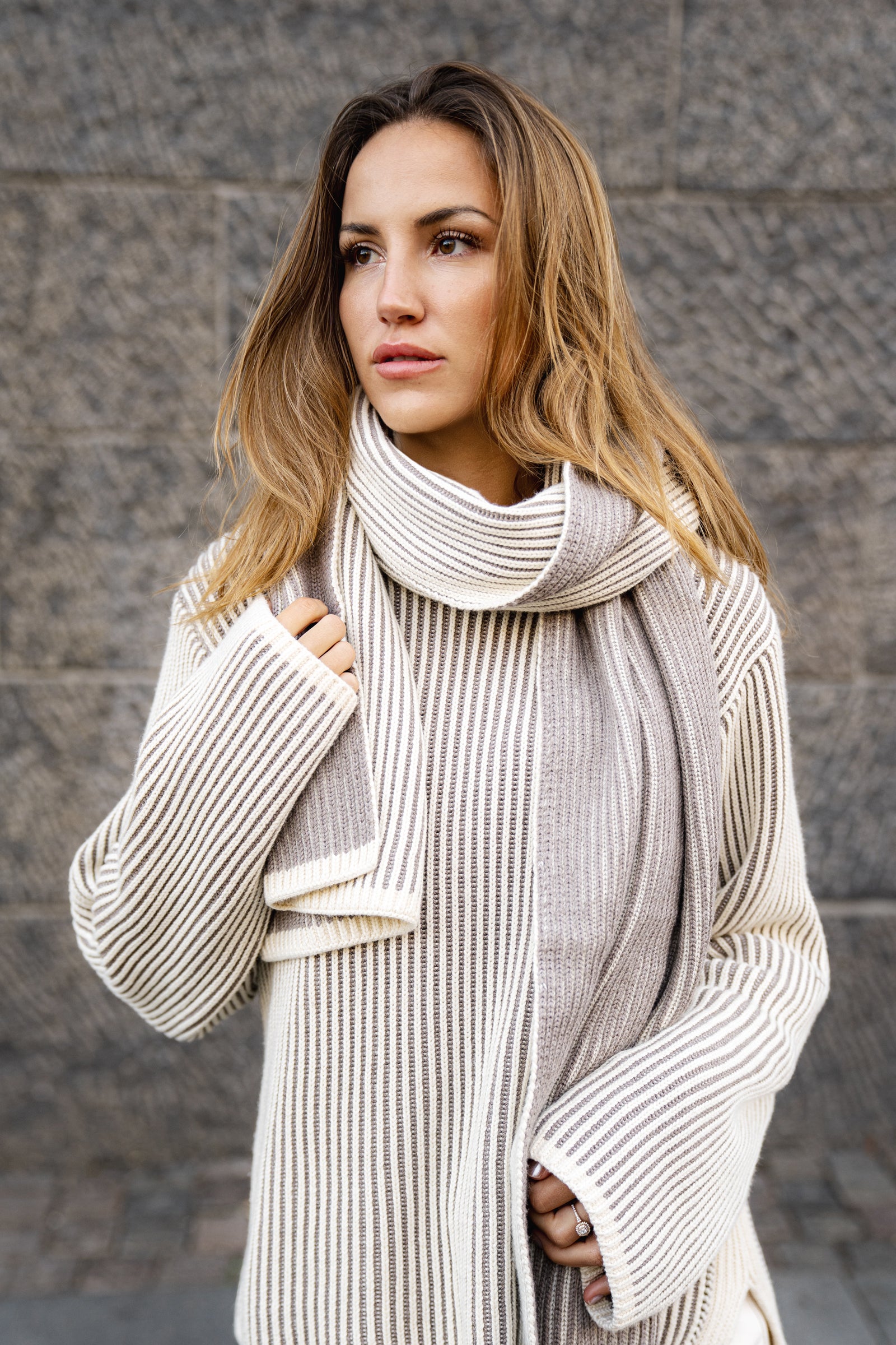 Woman wearing a long, light-colored scarf against a gray wall.