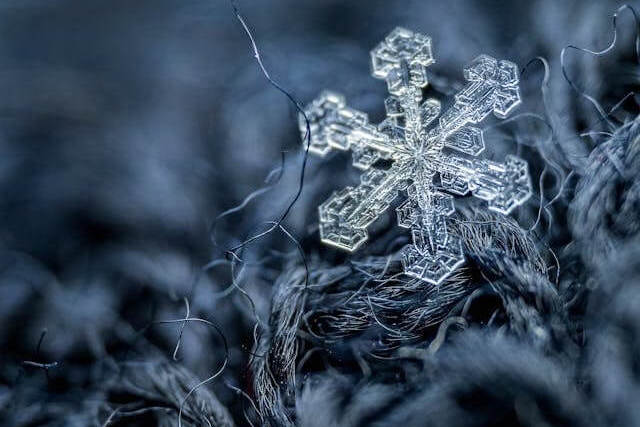 Close-up of a single intricate snowflake resting on dark, textured fabric fibers. The contrast highlights the delicate structure of the snowflake against the fabric's soft weave.