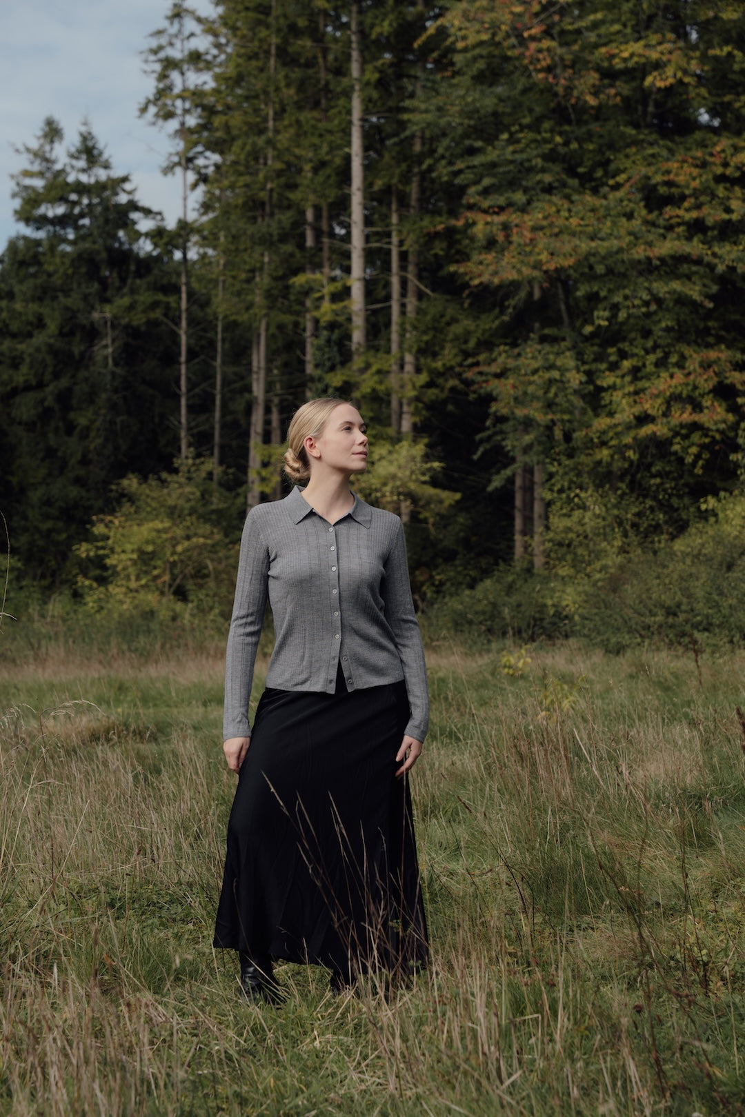 Grey Merino Polo Cardigan worn with a long black skirt in a grassy clearing.