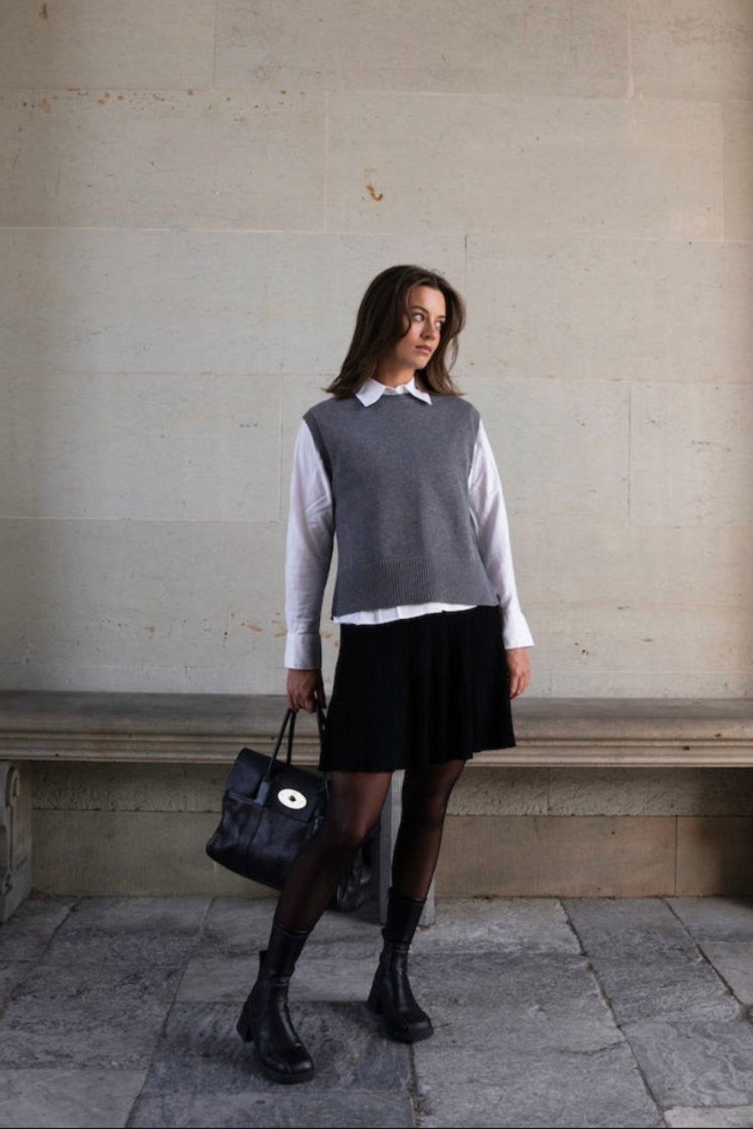 A woman stands in a stone archway corridor wearing a gray knit sweater vest over a white collared shirt, a black pleated skirt, black tights, and black ankle boots, while holding a black handbag.