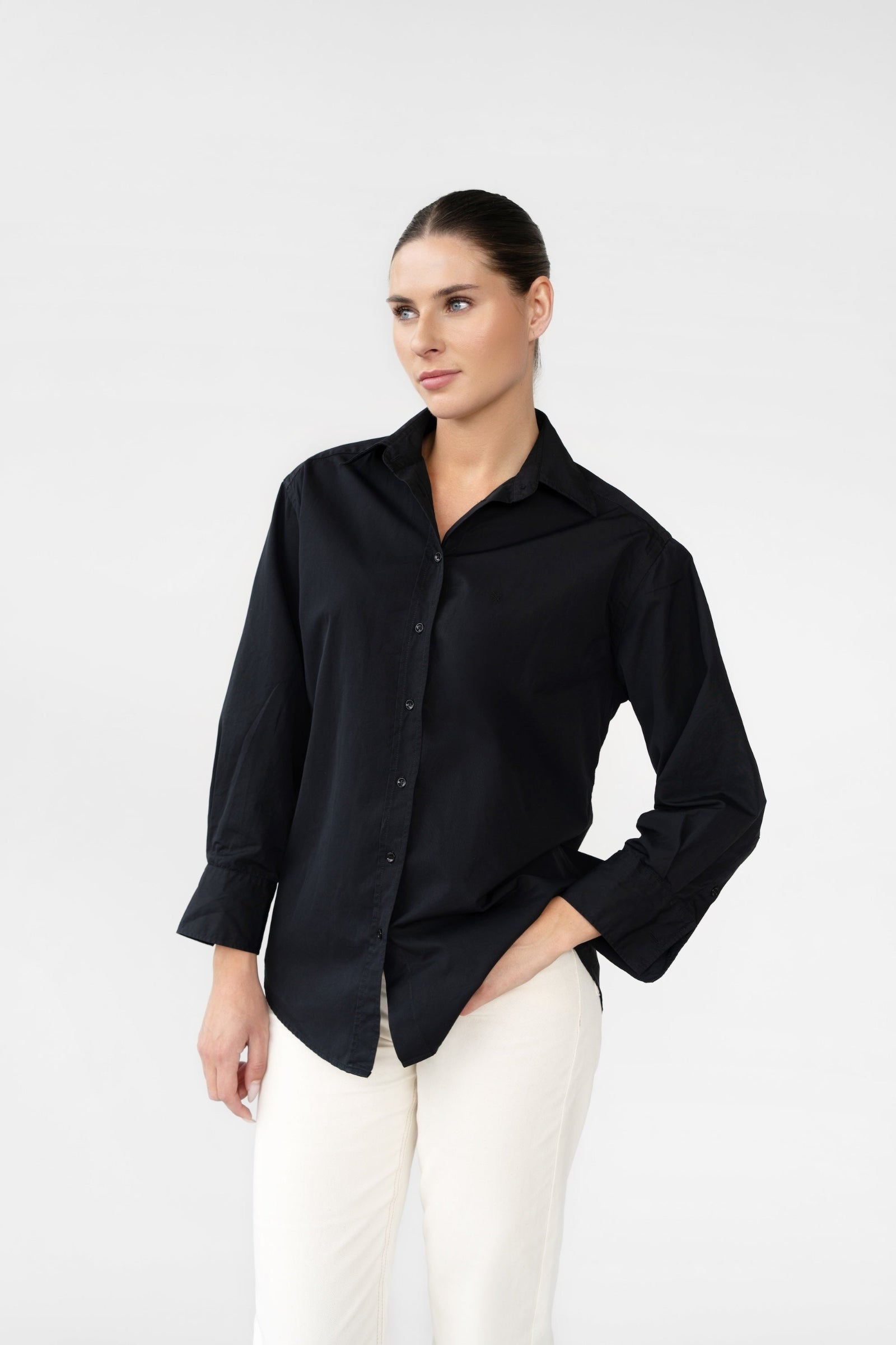 Front view of the CYME Supima Shirt in black, worn on-model half-tucked into off-white trousers. The relaxed crisp cotton shirt has a pointed collar, button placket and wide cuffs, shot in the studio against a light grey background.