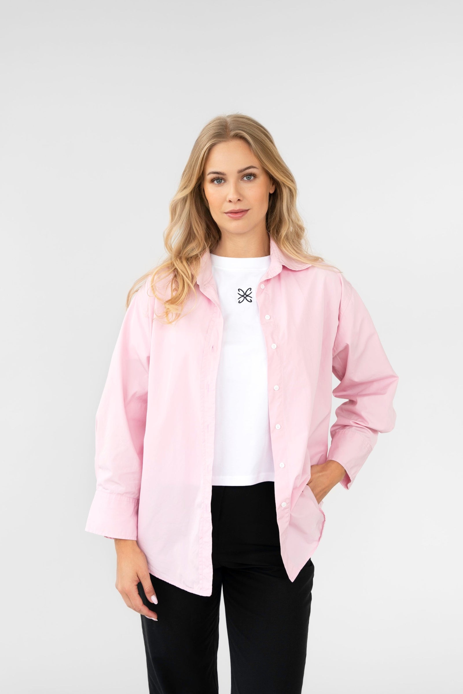 Woman wearing a light pink shirt over a white top with a black bow, standing against a plain background.