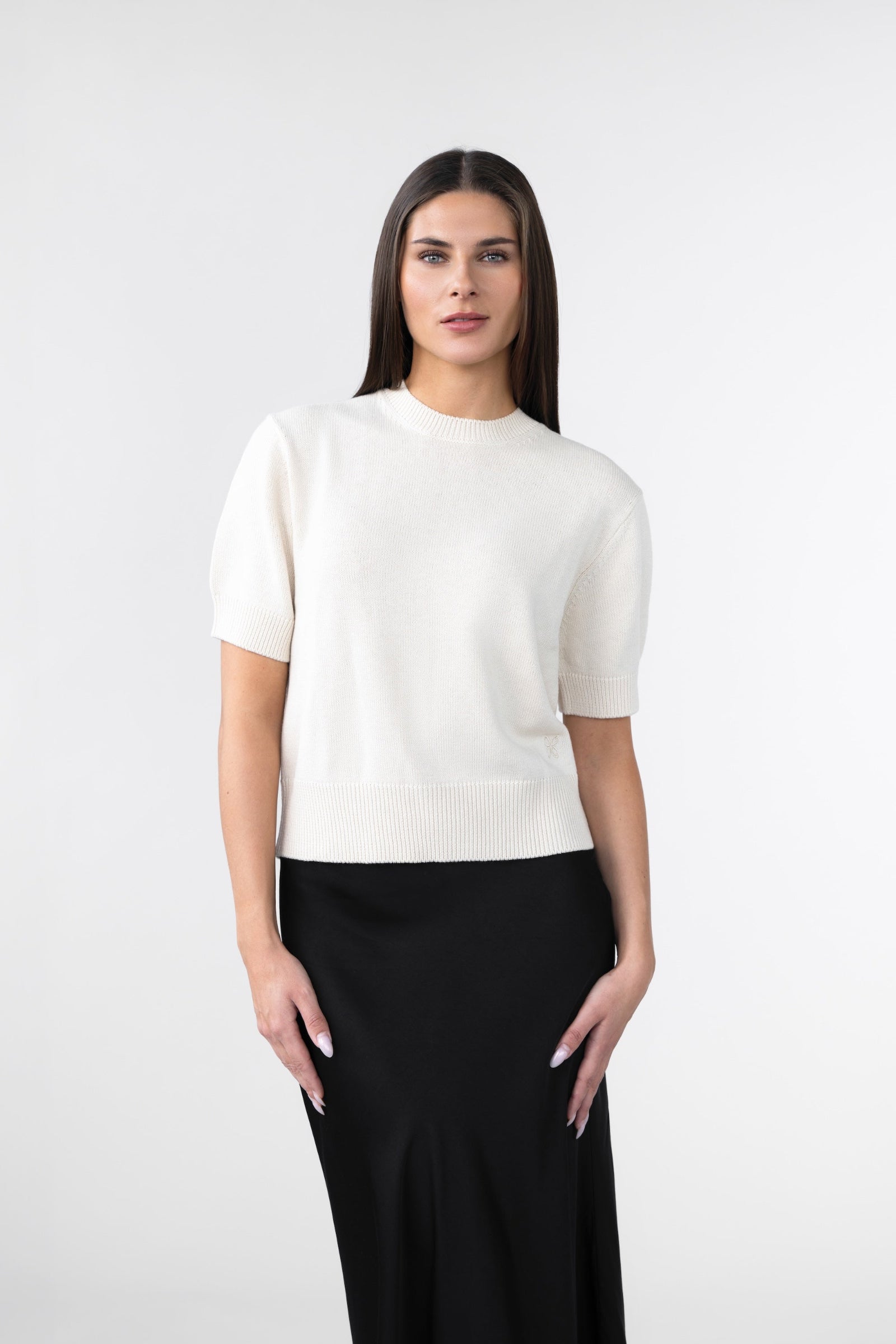 A woman wearing a white knitted tee and a black satin skirt against a gray background.