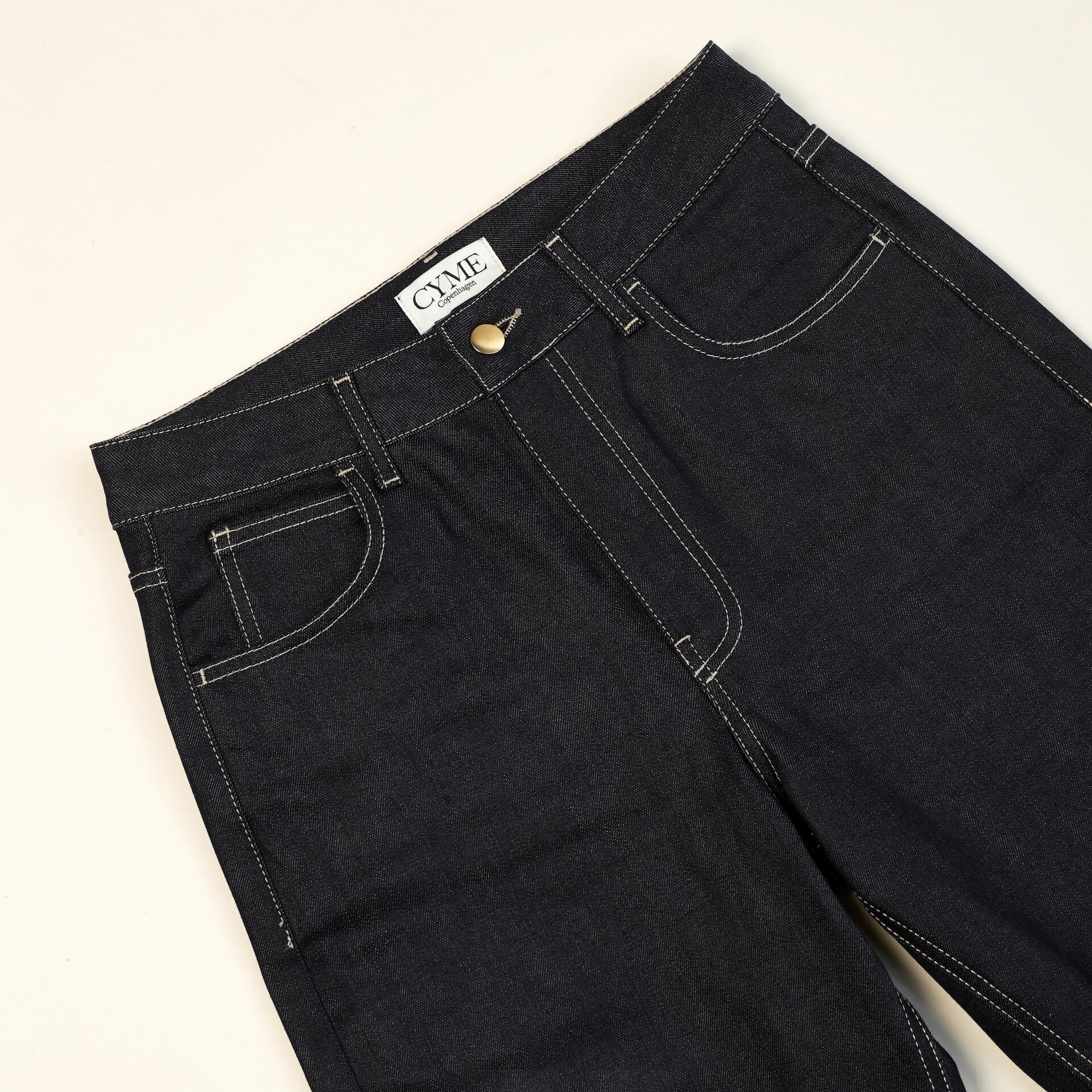 Close-up of a pair of dark blue high-waisted jeans from CYME Copenhagen. The jeans feature a classic five-pocket design with contrasting white stitching and a brass button closure. The CYME Copenhagen label is prominently displayed on the inner waistband.